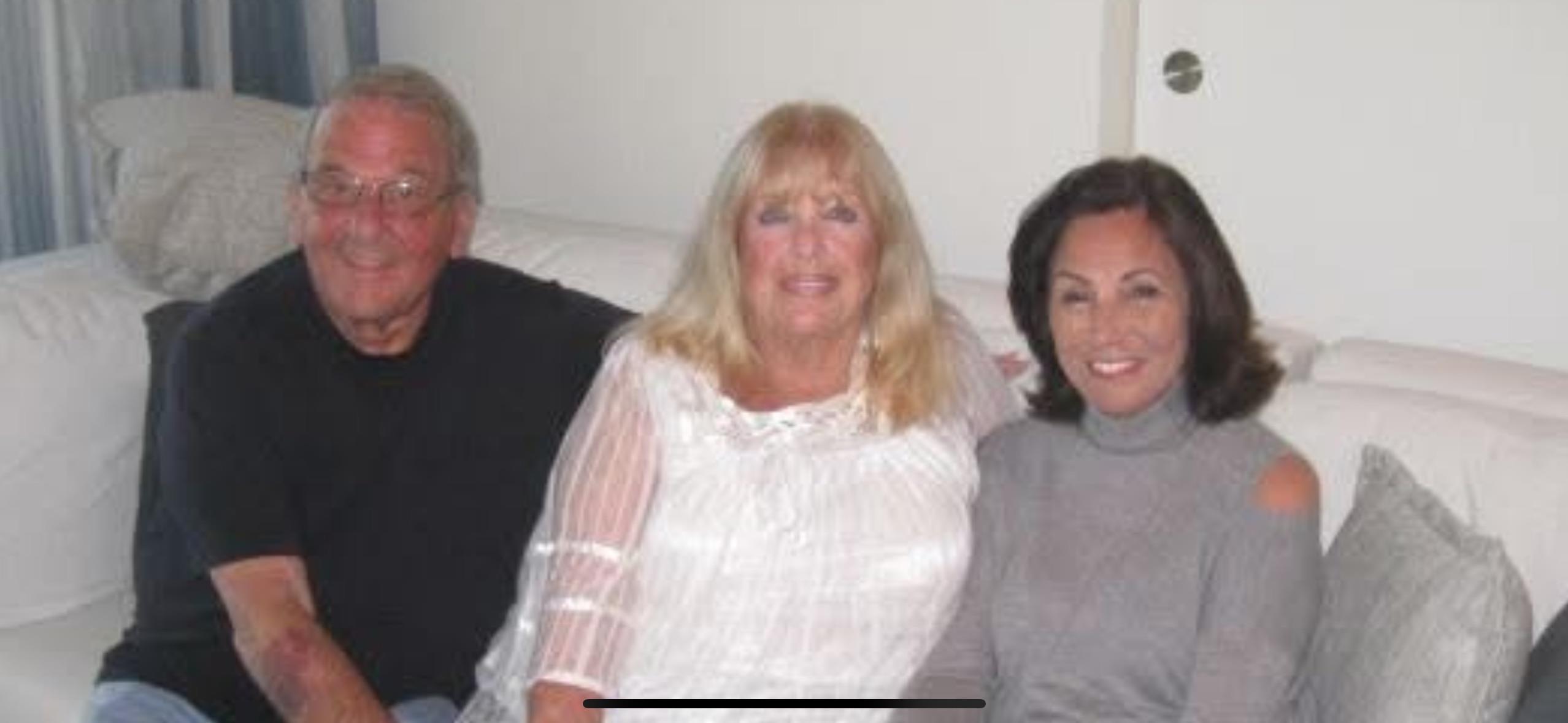 Three friends smile together on a comfortable couch in a well-lit living room setting.