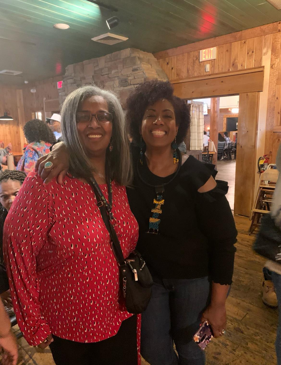 Two women smile broadly, embracing each other in a welcoming indoor setting filled with friends.