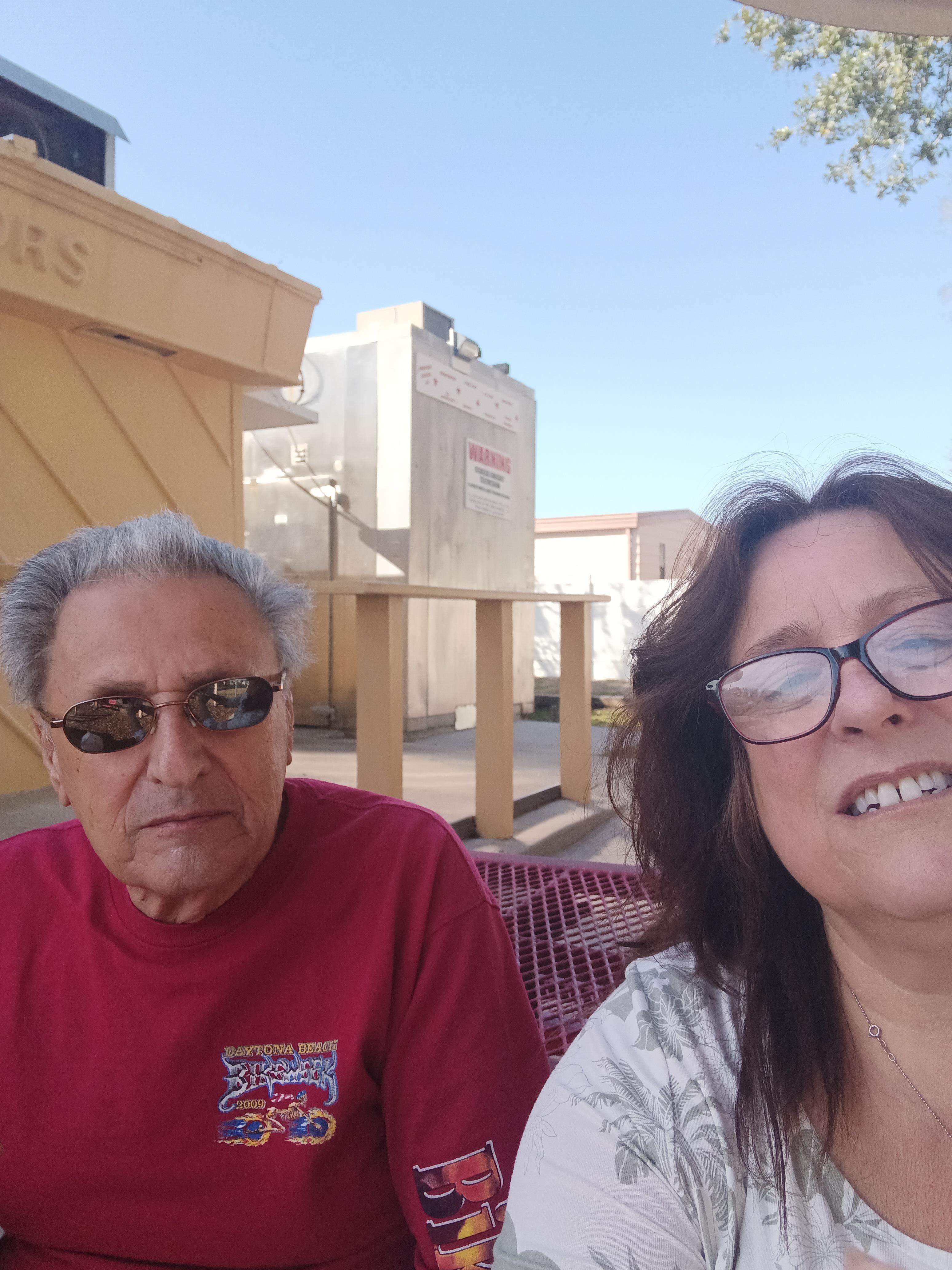 Two individuals are smiling and taking a selfie at a vibrant outdoor location under the sun.