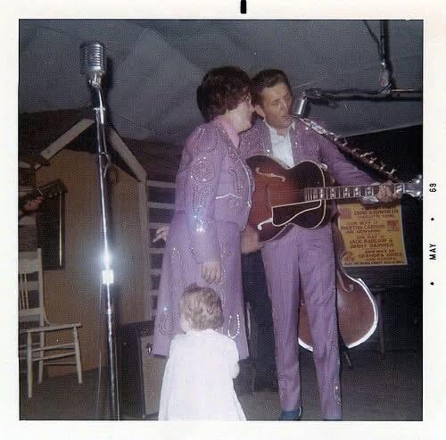 A couple in purple outfits sings on stage as a captivated child watches.