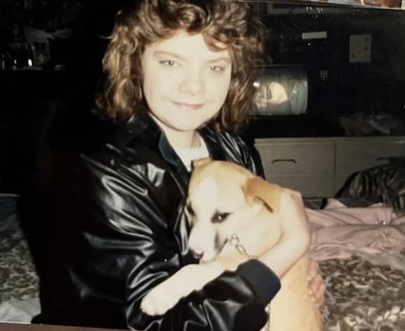 A young woman with curly hair sits on a bed, smiling while cuddling a dog in a warm environment.