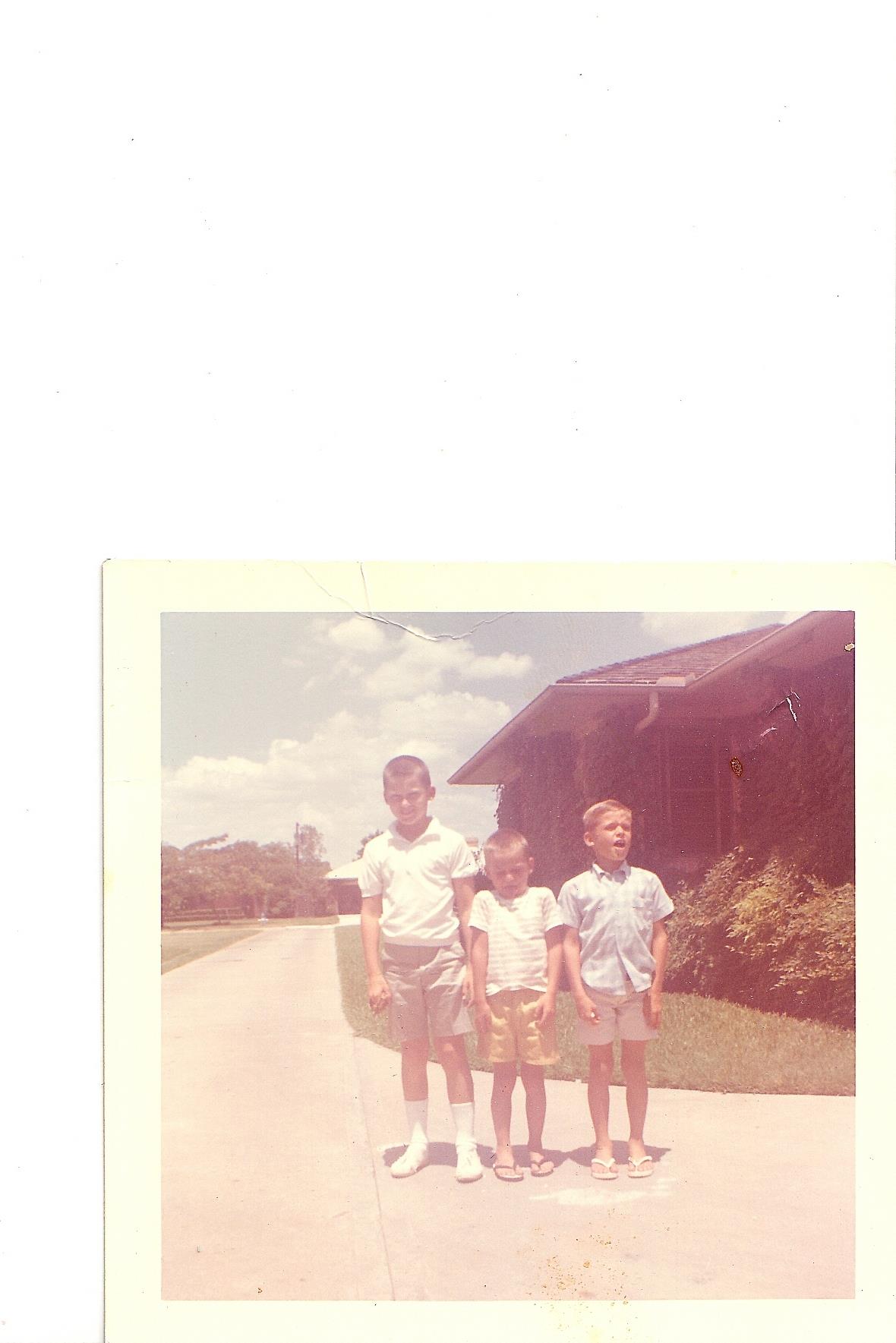Three young boys stand on a driveway in a residential area under a clear sky.
