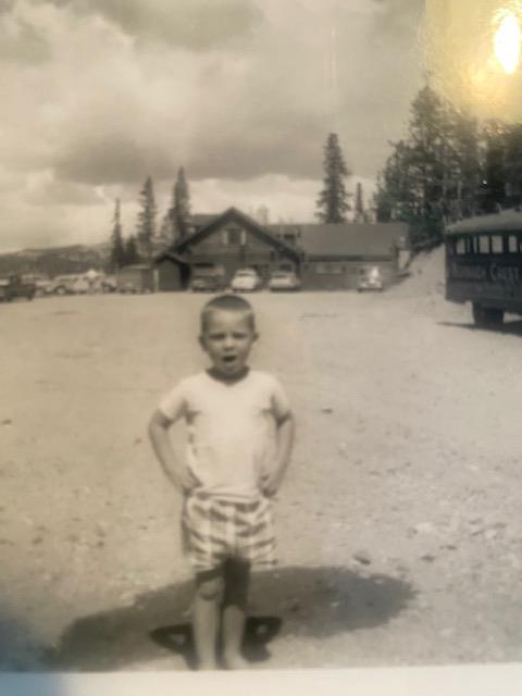 A boy stands on a gravel road, hands on hips, by trees and a mountain lodge.