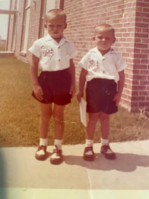 Two children dressed in identical clothing pose for a photo on a sidewalk near a school.