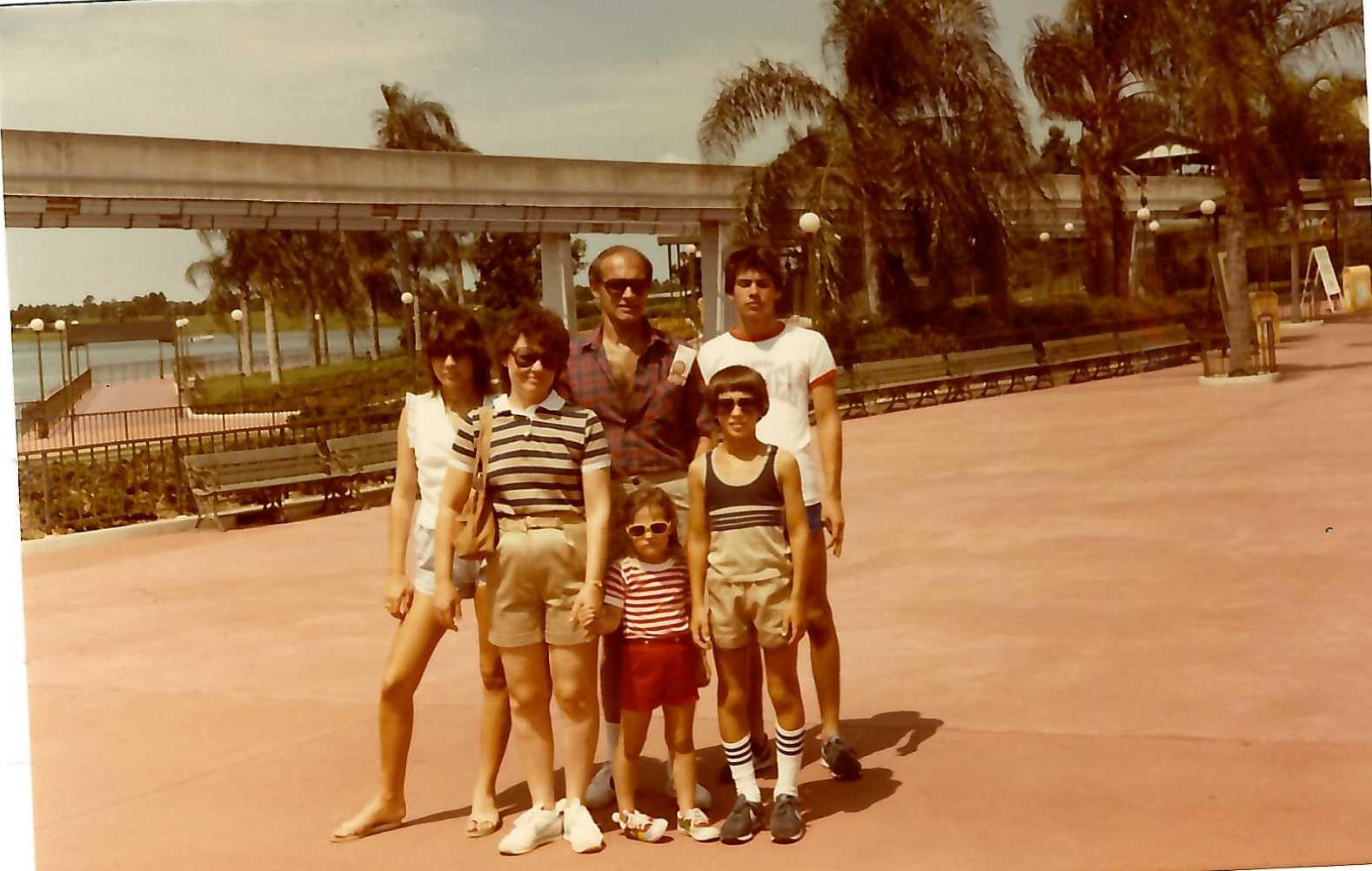 Six family members stand together in a park filled with palm trees on a sunny day.