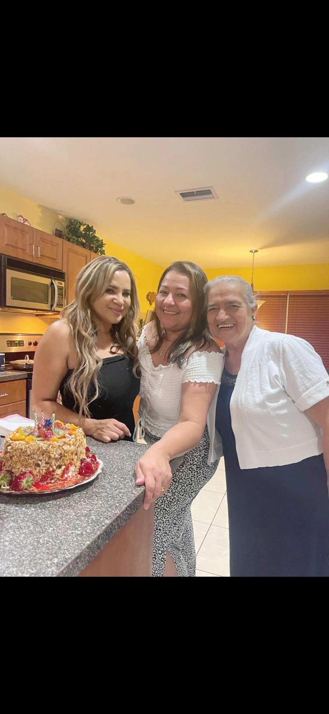 Three women smile together in a cheerful kitchen while celebrating with a decorated cake.