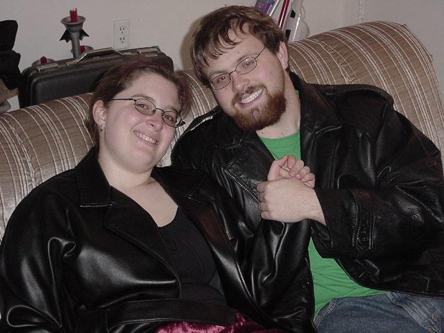 A couple in matching leather jackets sits on a couch, smiling and holding hands.