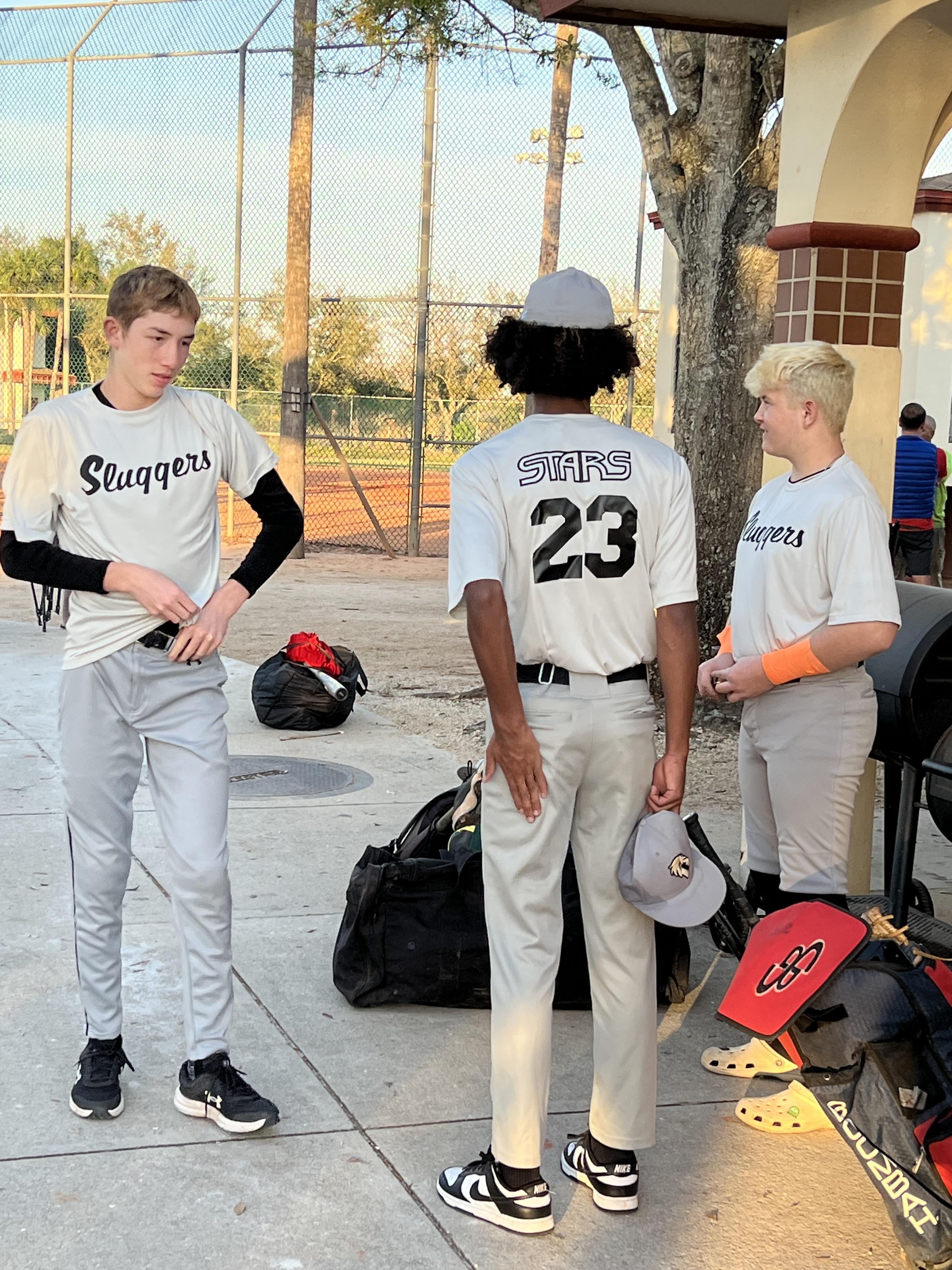 Three young players chat and adjust their gear before a baseball game at a local field.