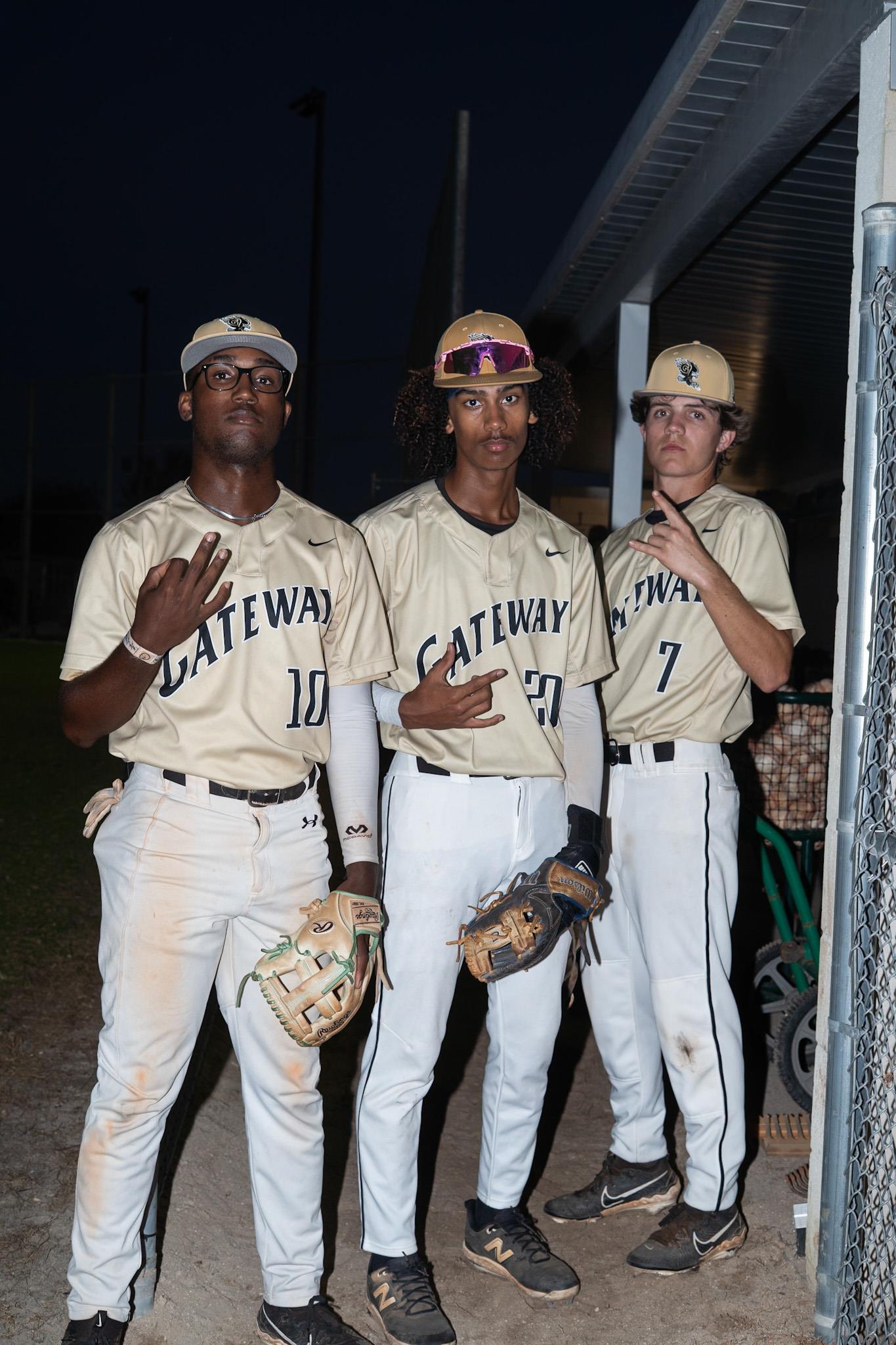 Three young athletes wearing beige baseball uniforms stand under a dugout during twilight.
