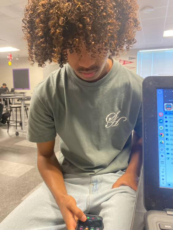 A young student with curly hair is absorbed in his phone while seated at a desk.