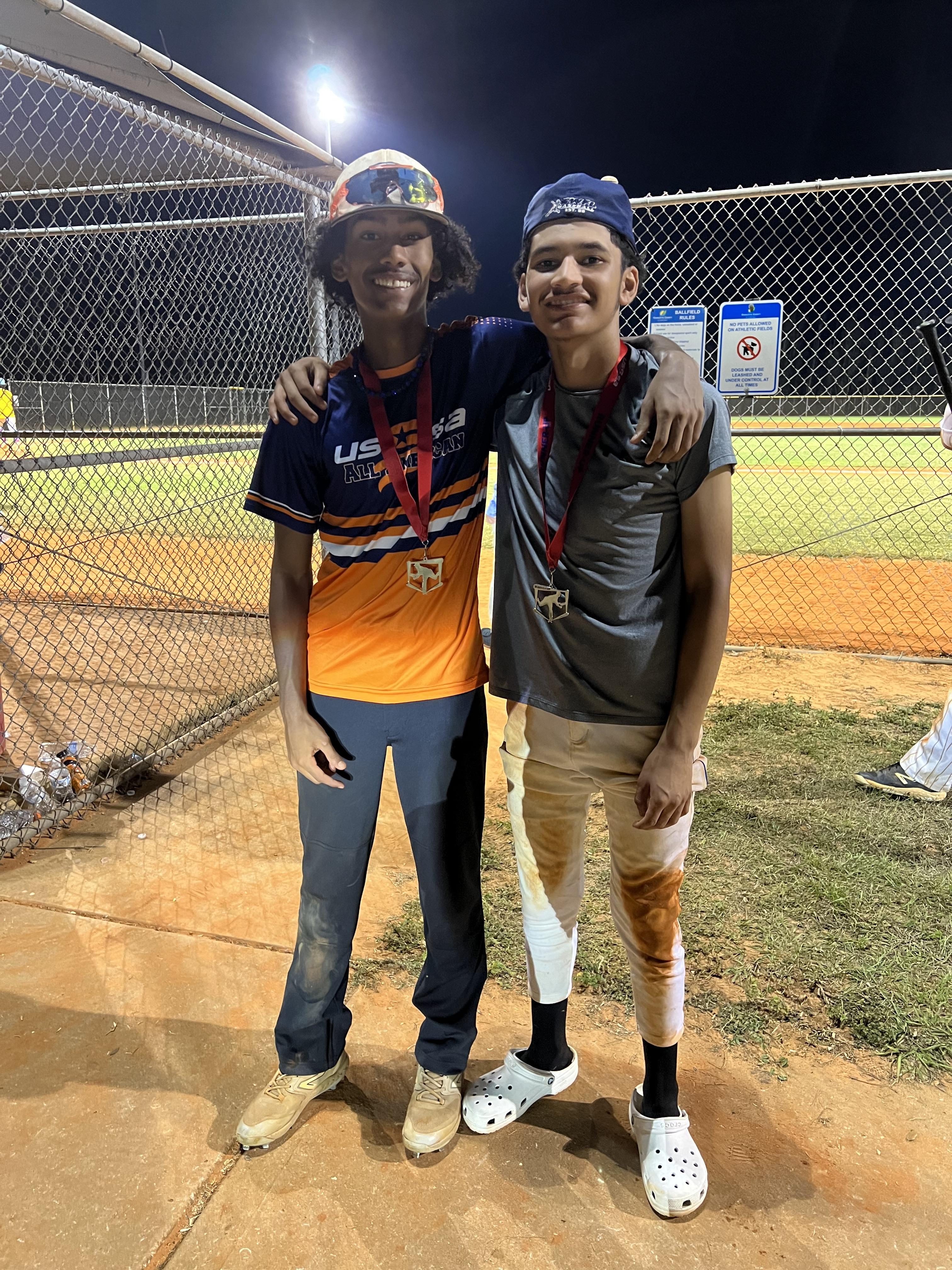 Two young baseball players smile proudly after winning a game under the stadium lights.