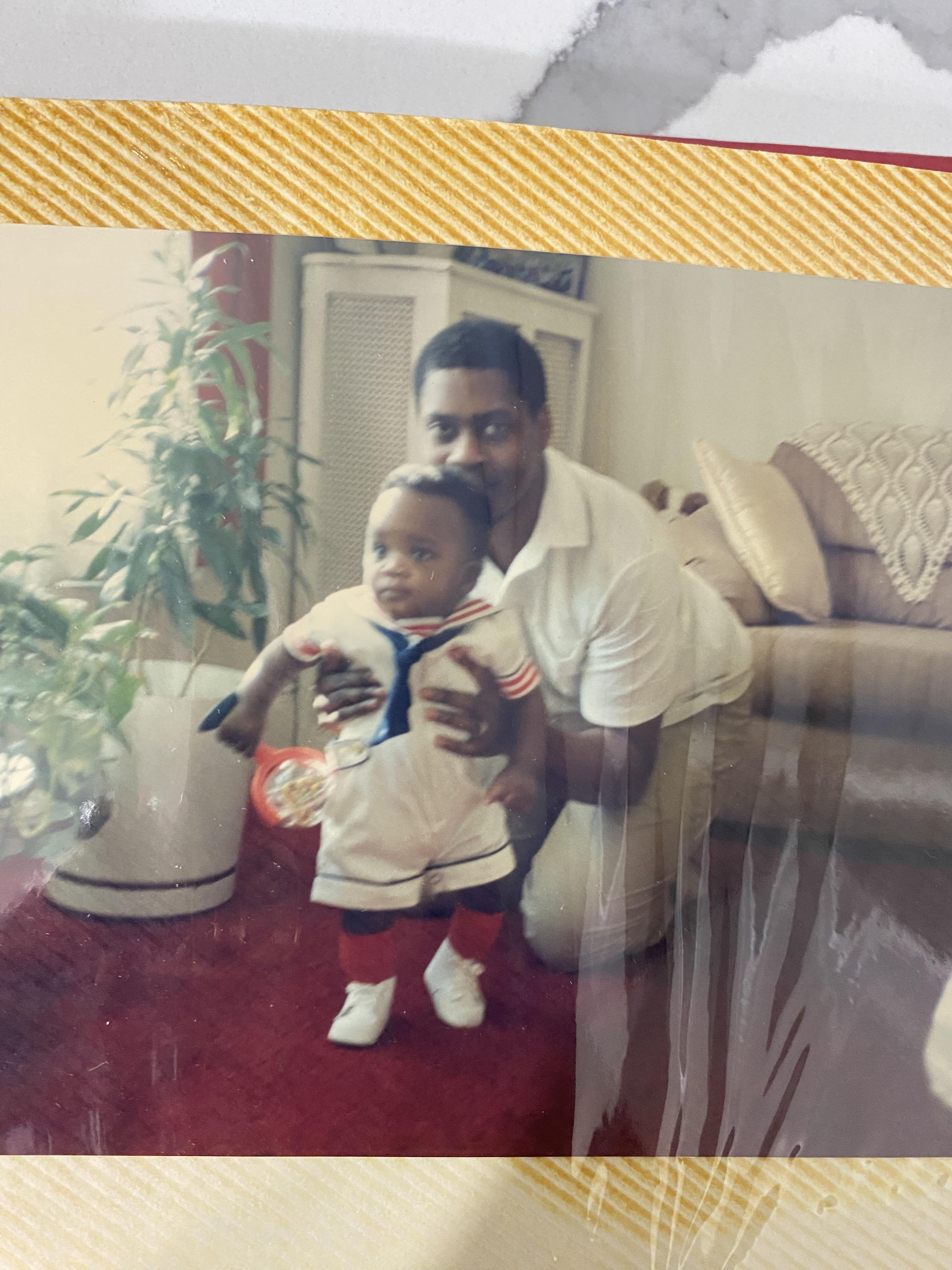 A man lifts his toddler son, both smiling in a bright living room filled with sunlight.