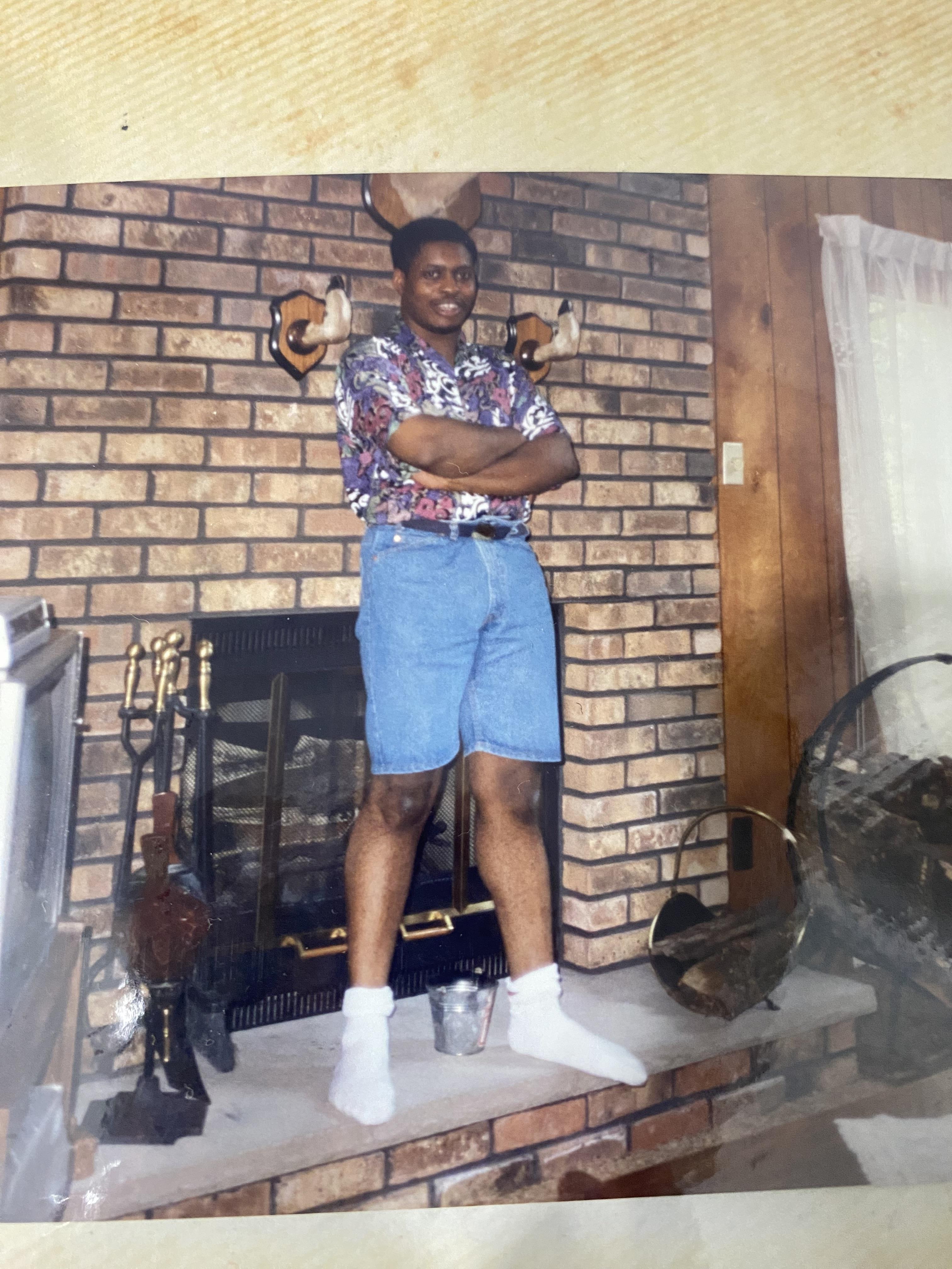 A man stands with crossed arms on a hearth in a cozy living room, wearing a floral shirt and shorts.