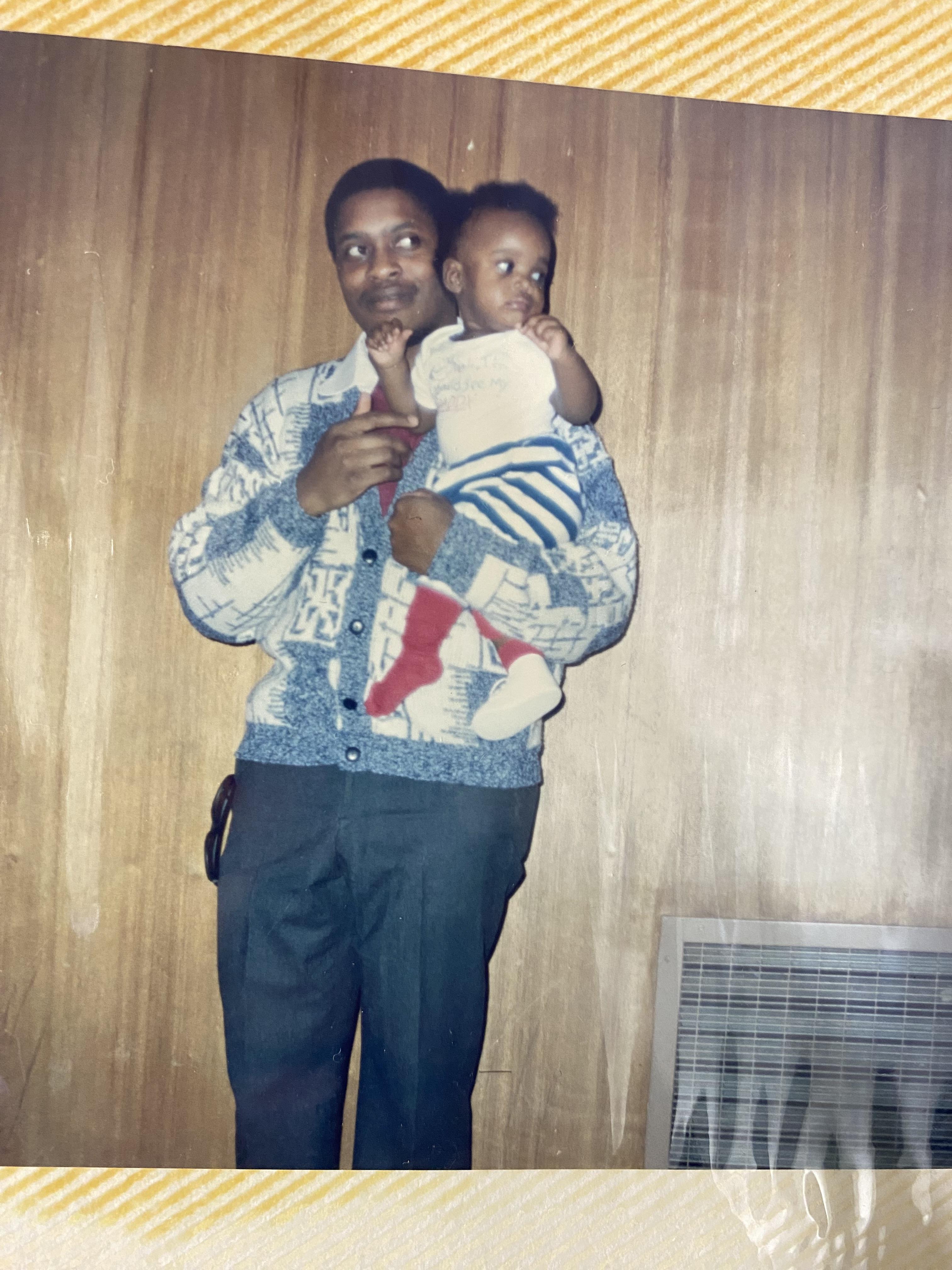 A father holds his young child in a warm indoor space with wooden paneling, sharing a tender moment.