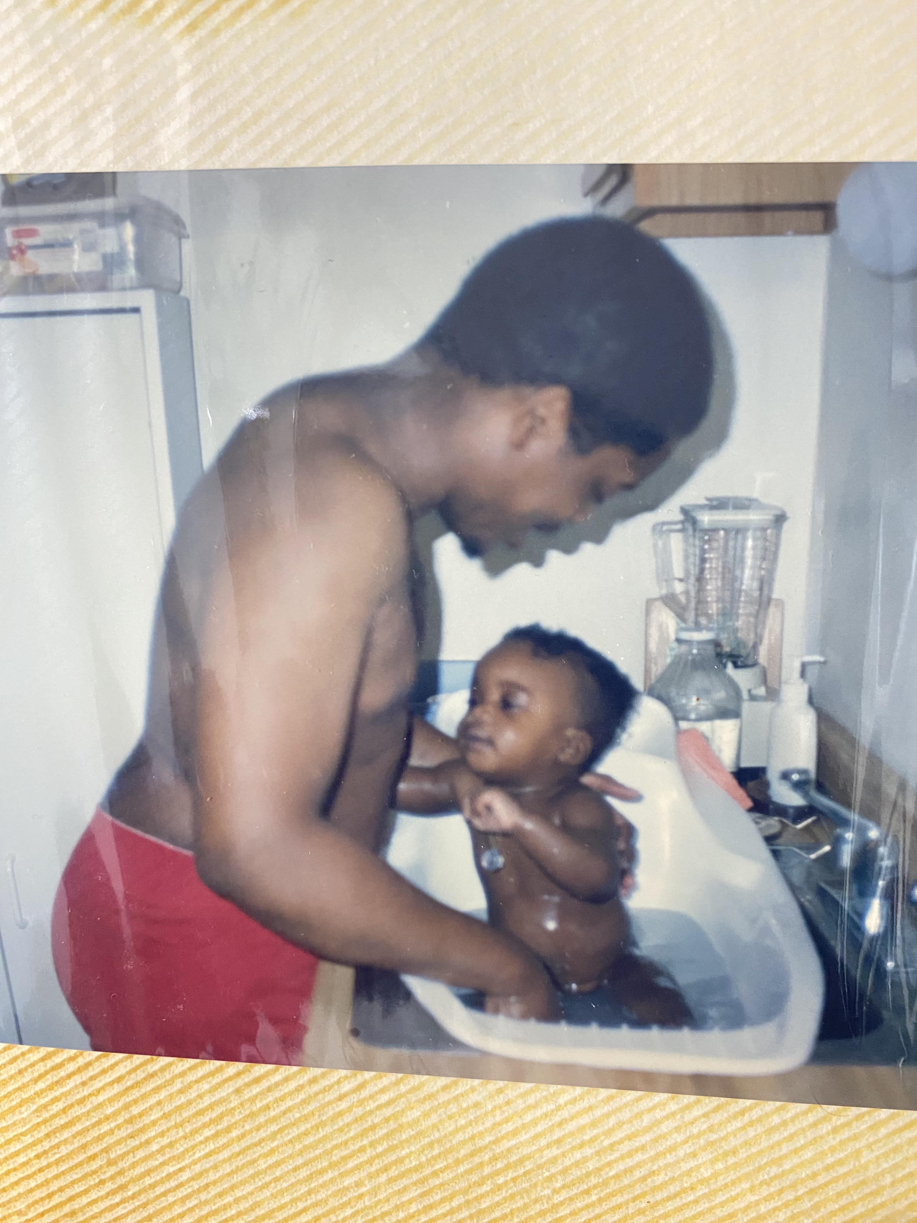 A father joyfully interacts with his baby while giving them a bath in a kitchen sink.