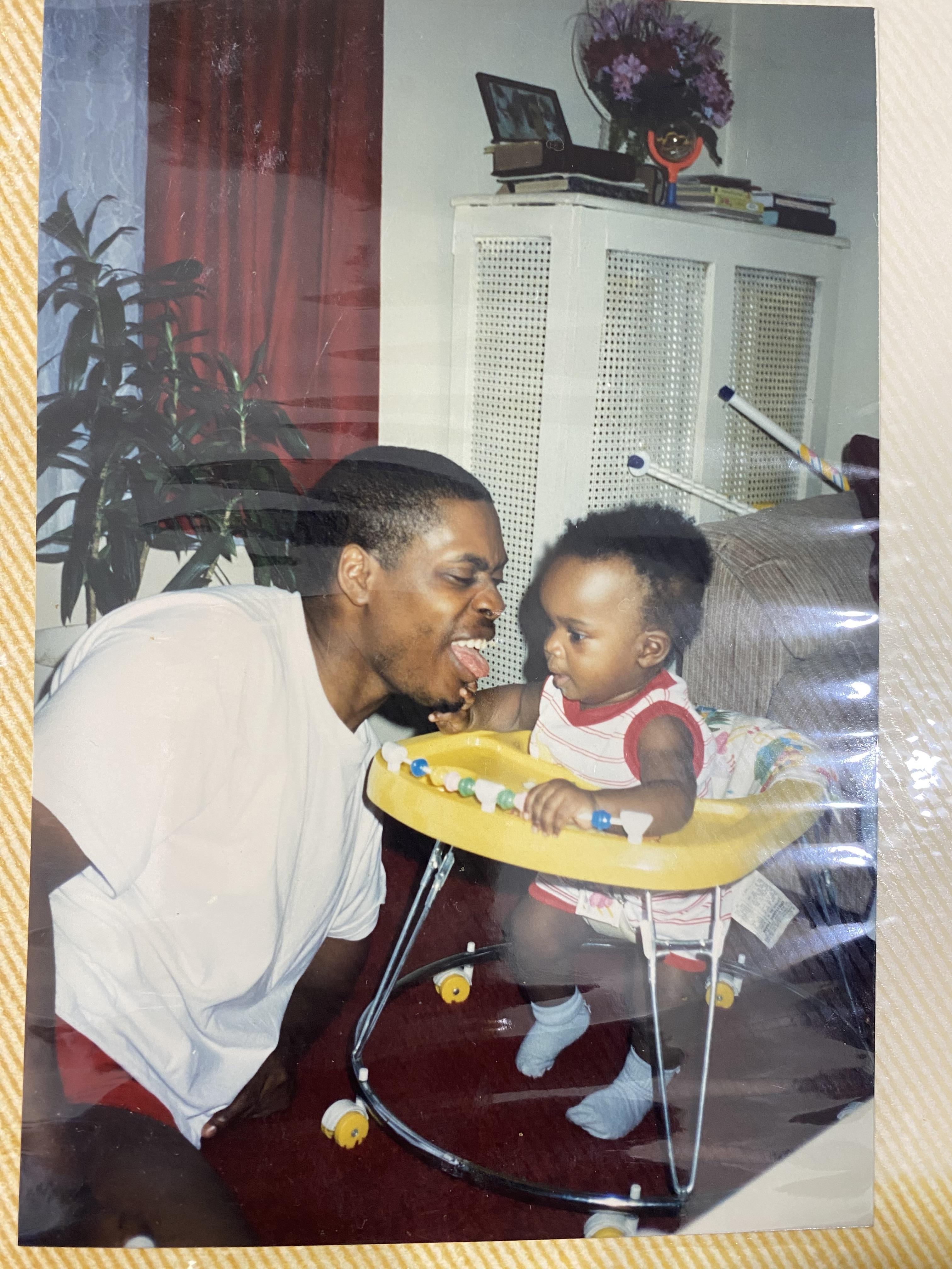 A father interacts playfully with his baby, who is in a colorful walker, in their home.