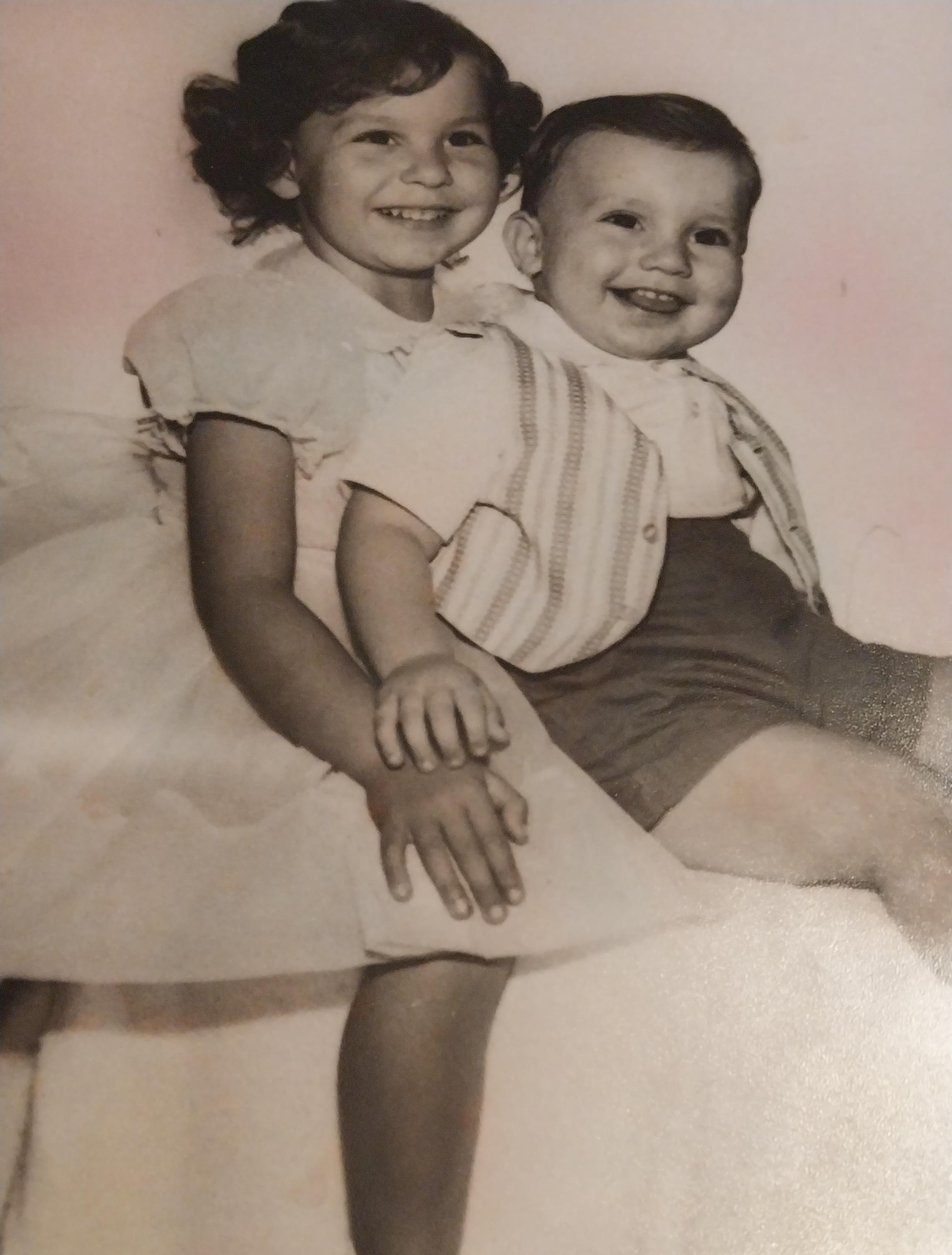 Two children share a joyful moment, sitting closely and smiling while dressed in vintage clothing.