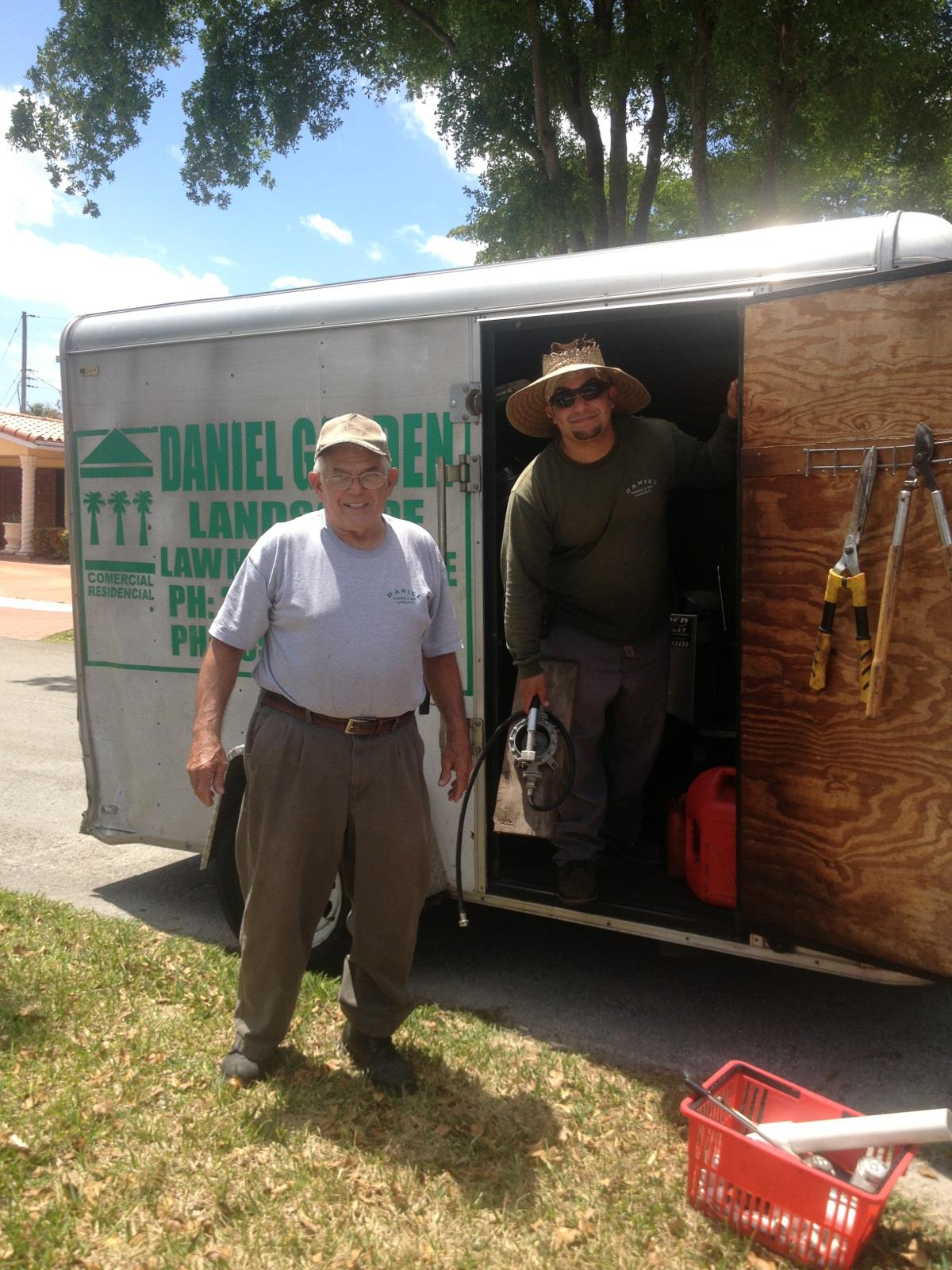 Two professionals prepare for landscaping tasks outside their trailer in daylight.
