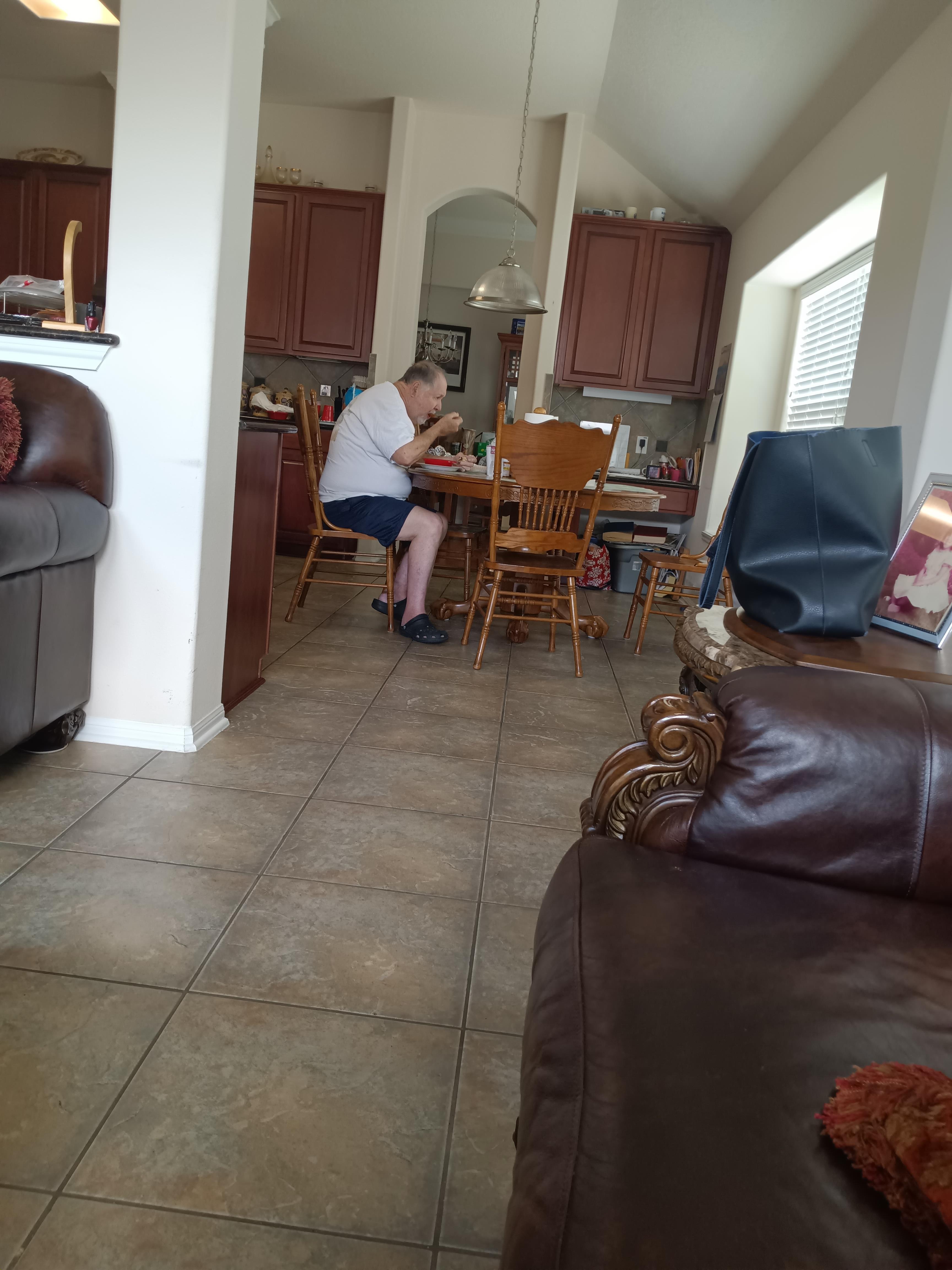 An older man is contentedly eating at a wooden table in a well-lit kitchen area.