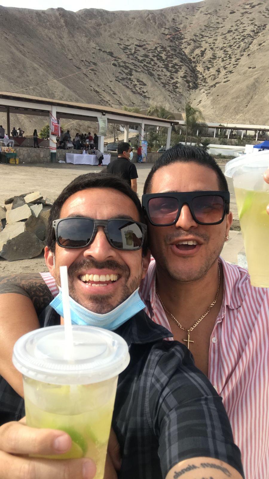 Two friends smile joyfully while raising their drinks at an outdoor event near the beach.