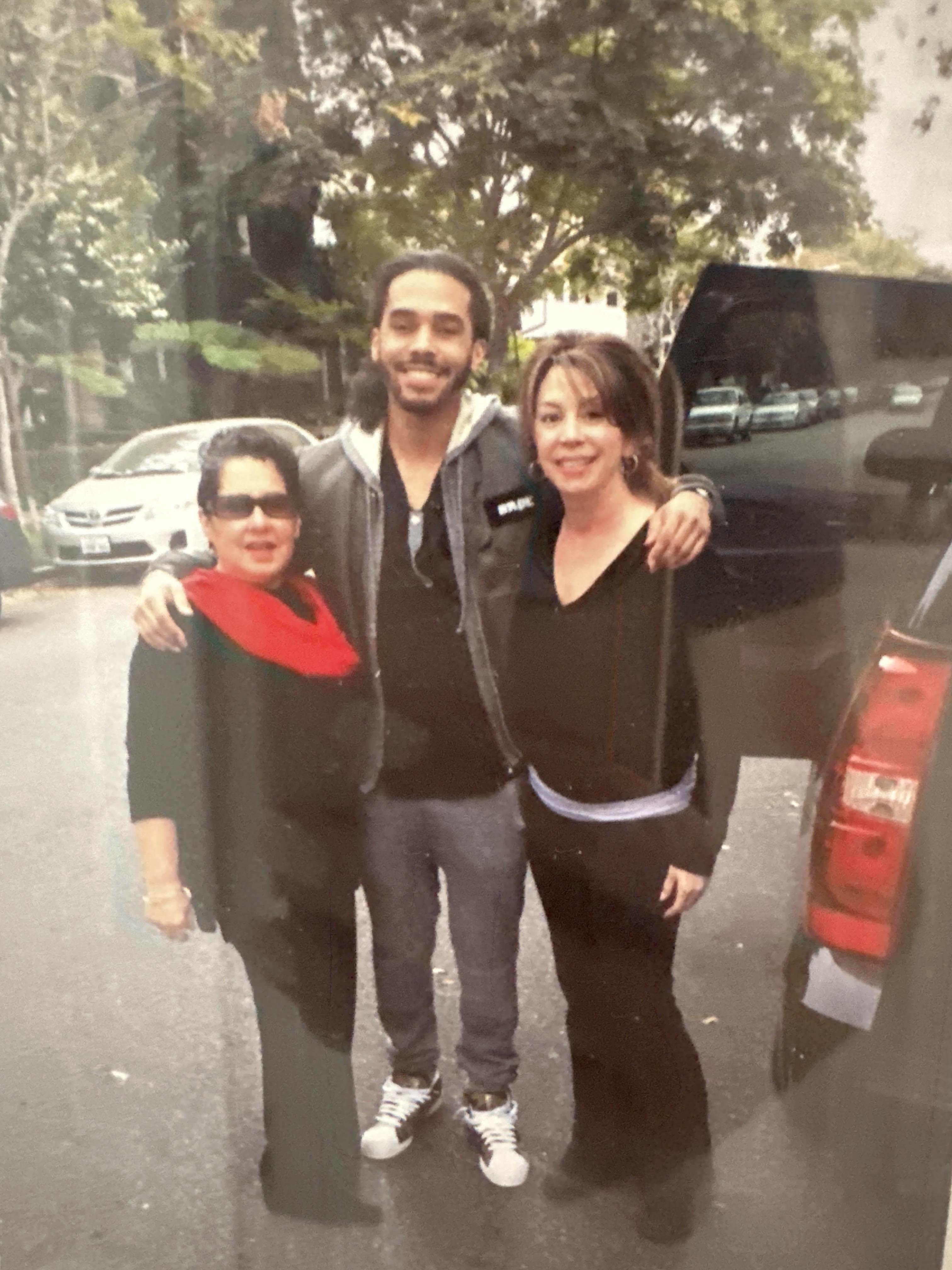 Two women stand on either side of a smiling man, all enjoying a sunny day in a lively neighborhood.