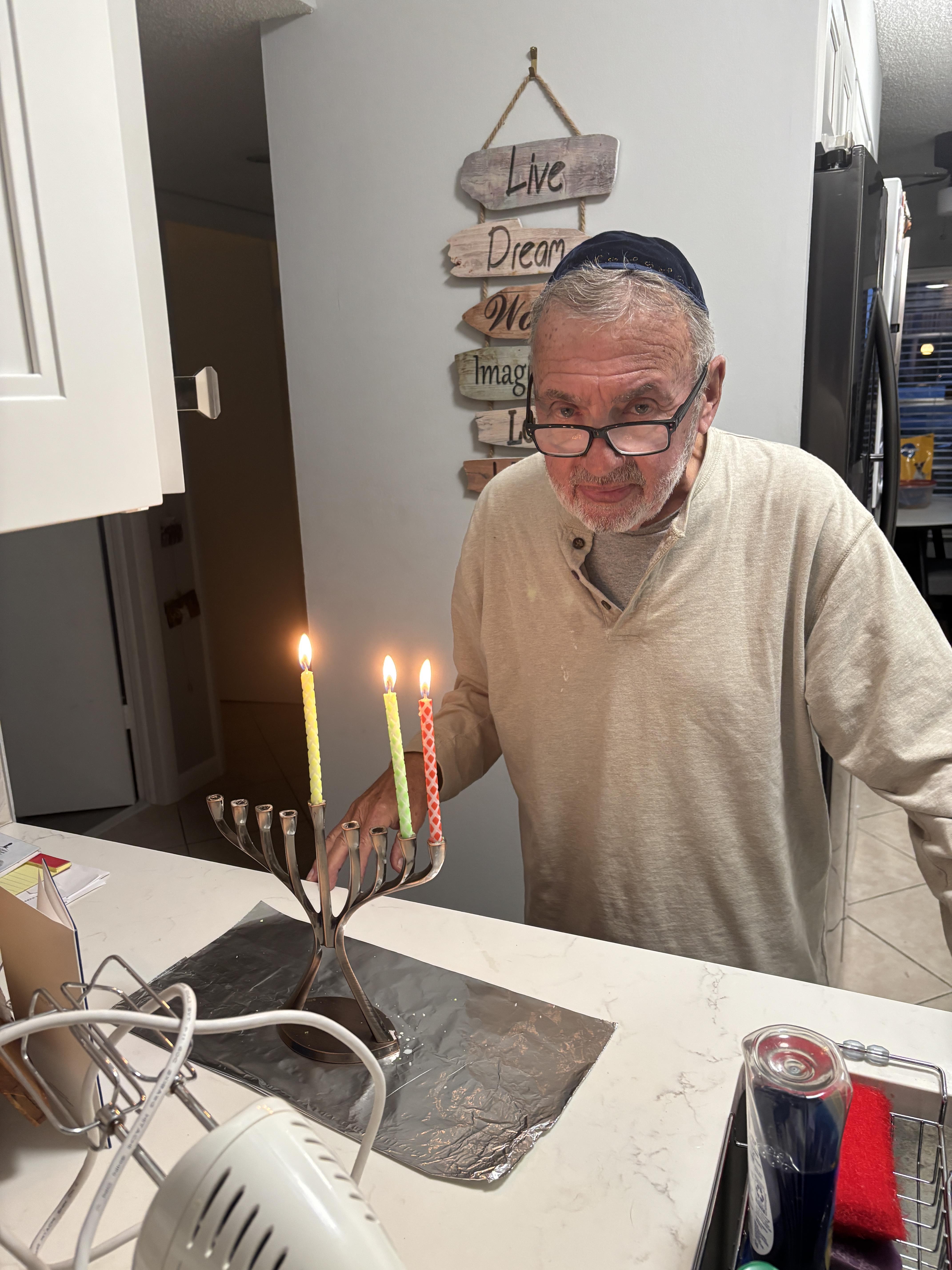 An elderly man prepares to light candles on a menorah in a warm kitchen setting at night.