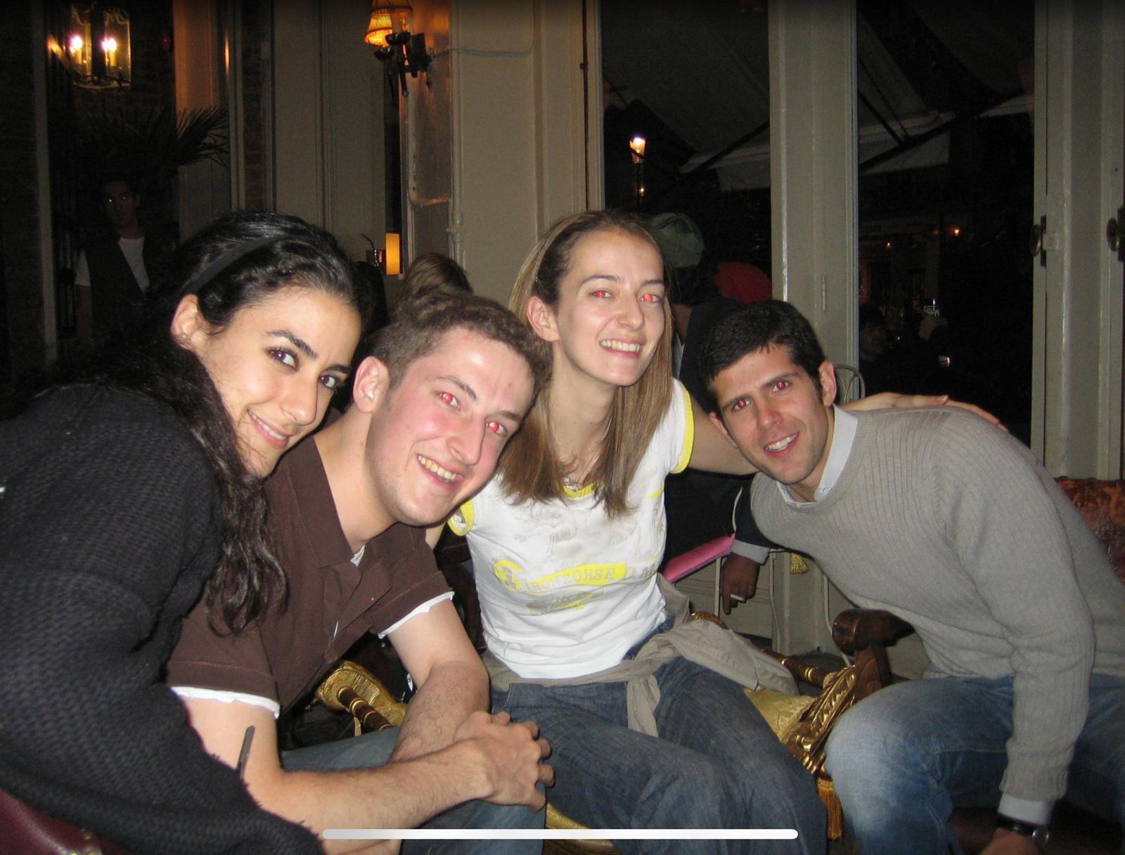 Four friends are smiling and posing together in a lively cafe setting during the evening.