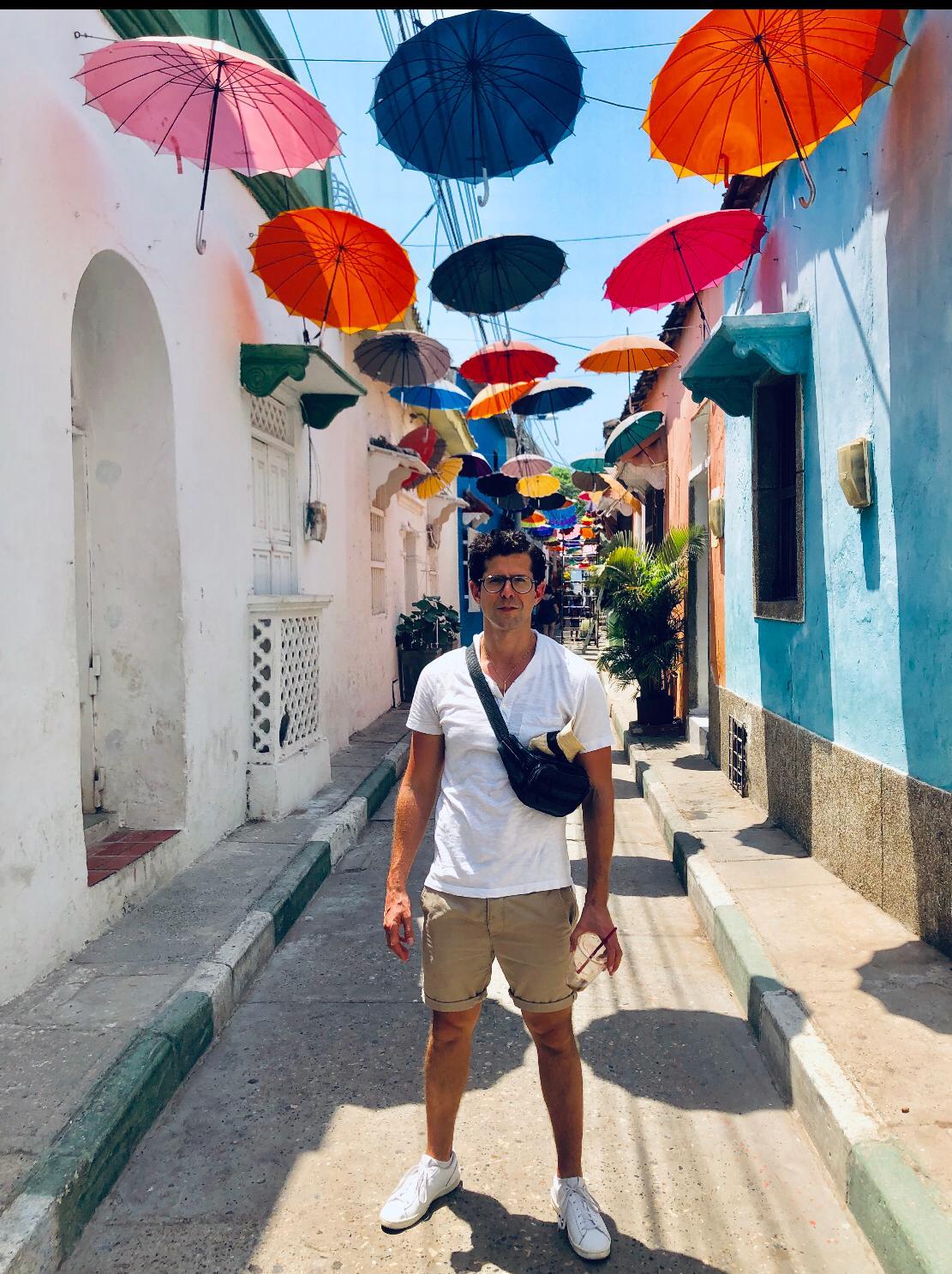 A person stands confidently in a narrow street adorned with colorful umbrellas overhead.