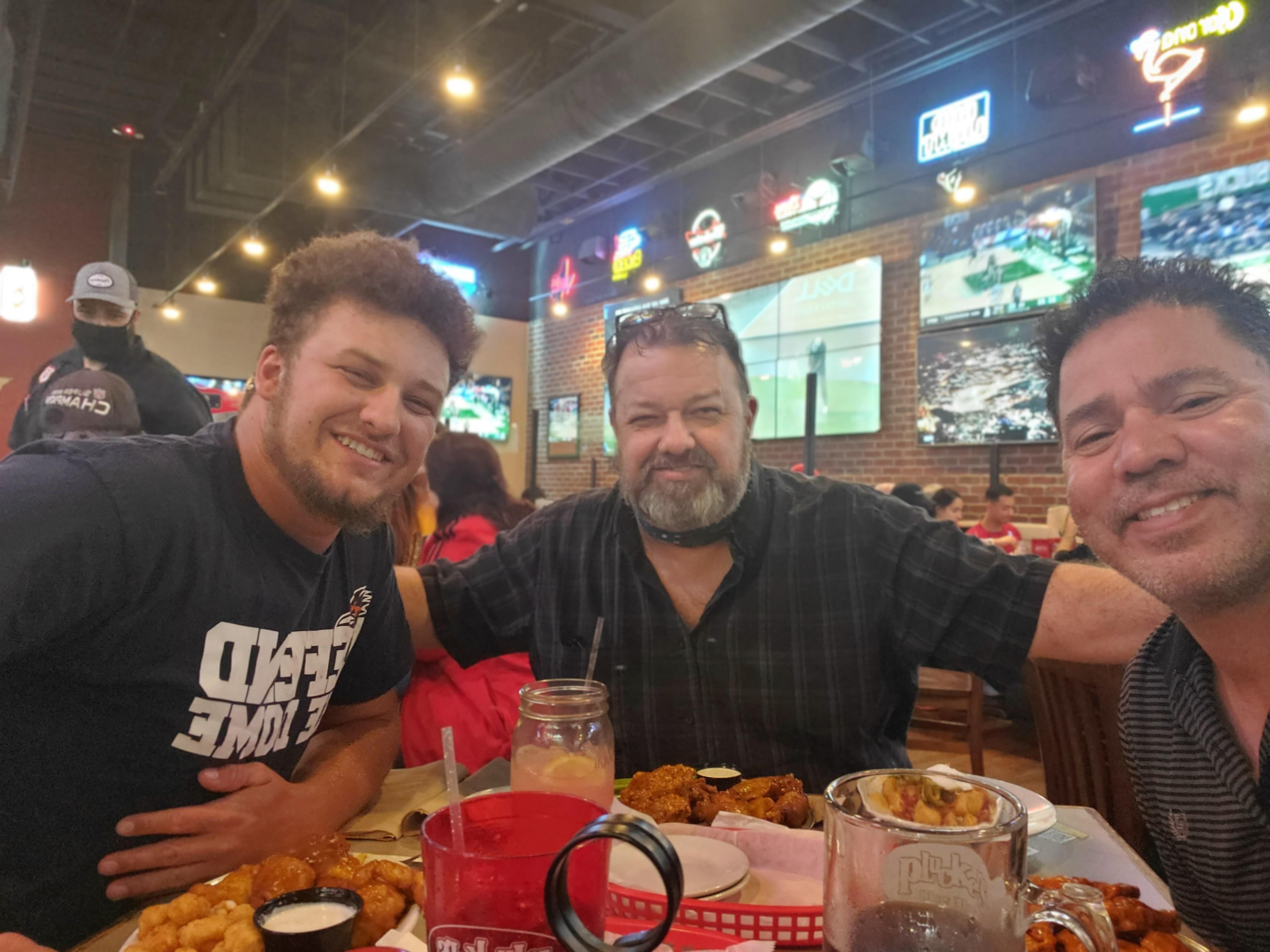 Three men share laughter and food at a vibrant restaurant filled with sports screens and fans.