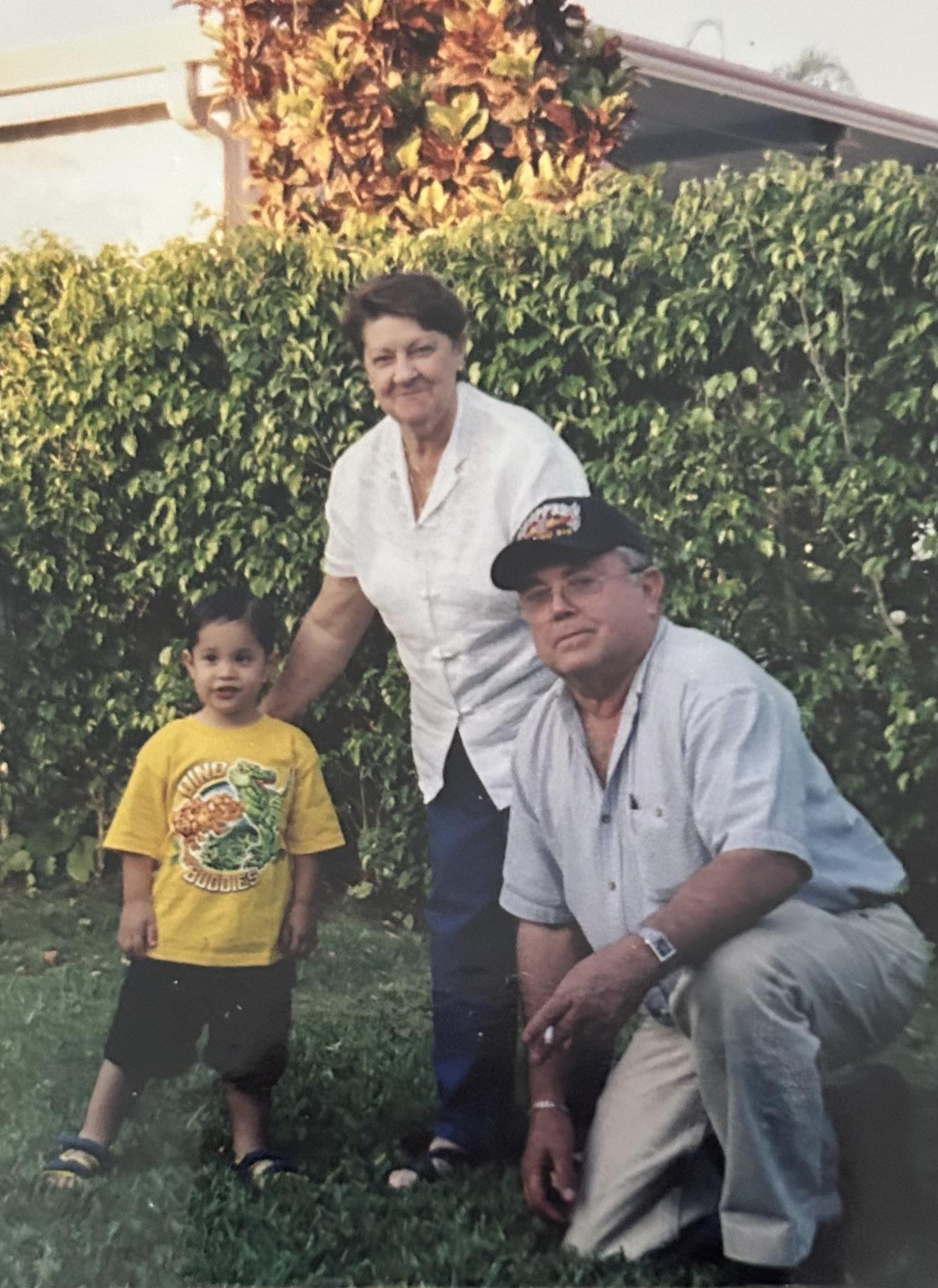 A young boy stands next to his smiling grandparents in a lush green garden.
