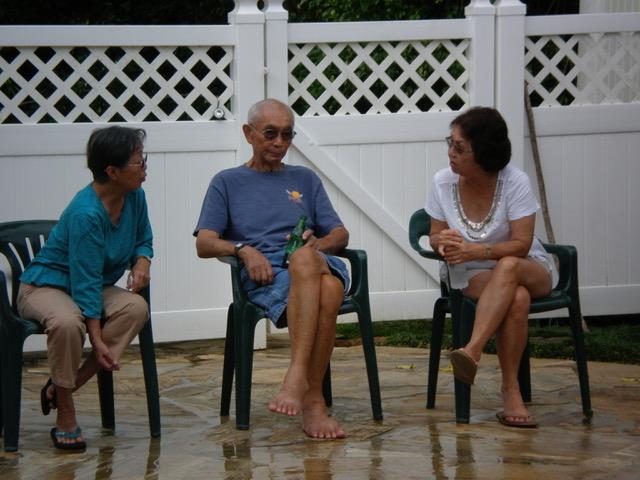 Three friends sit comfortably in chairs, relaxing and chatting near a pool in sunny weather.