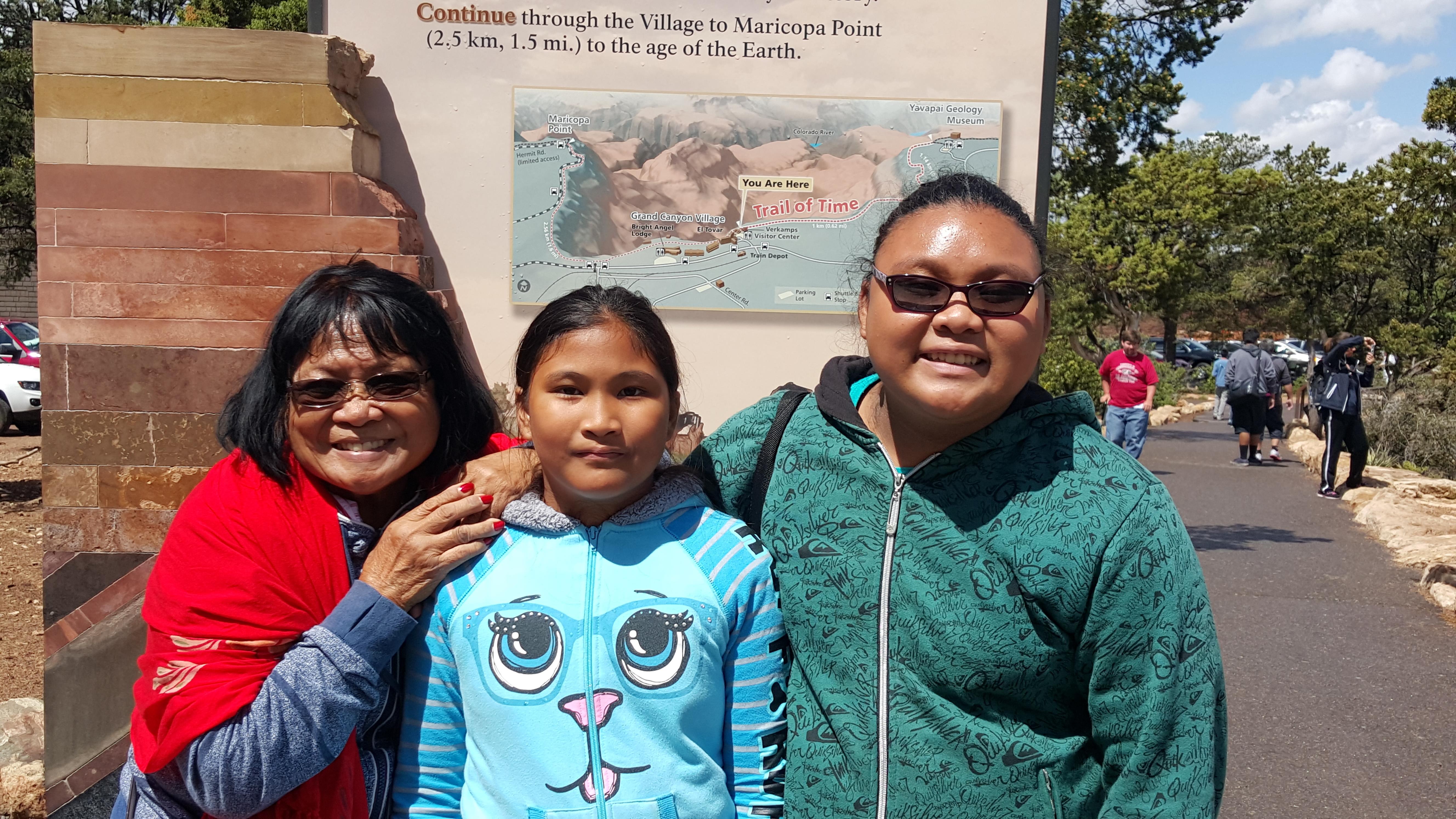An older woman, a middle-aged woman, and a young girl pose happily near a Grand Canyon sign.