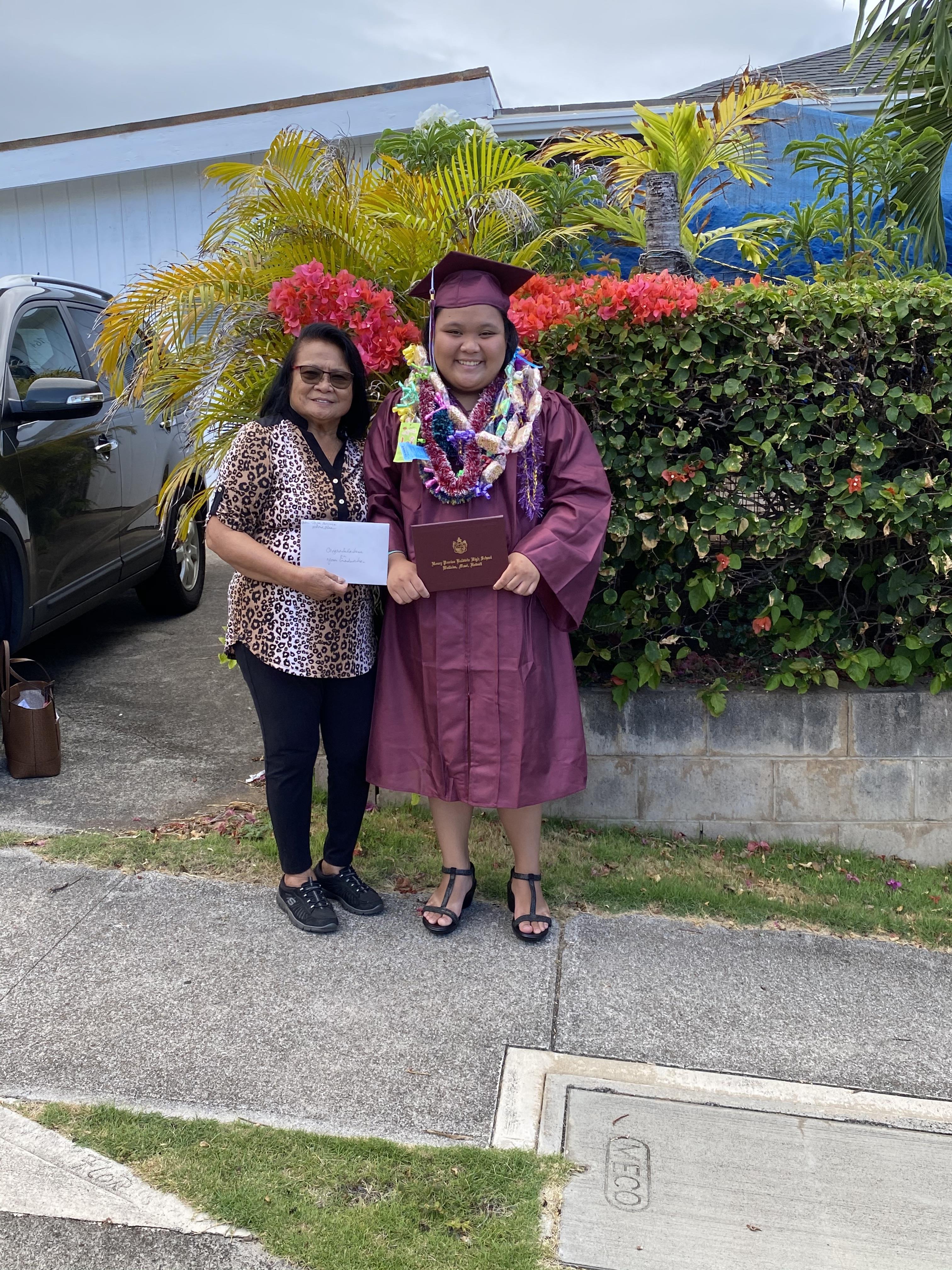 Graduate wearing cap and gown smiles while posing with loved one amidst colorful flowers.