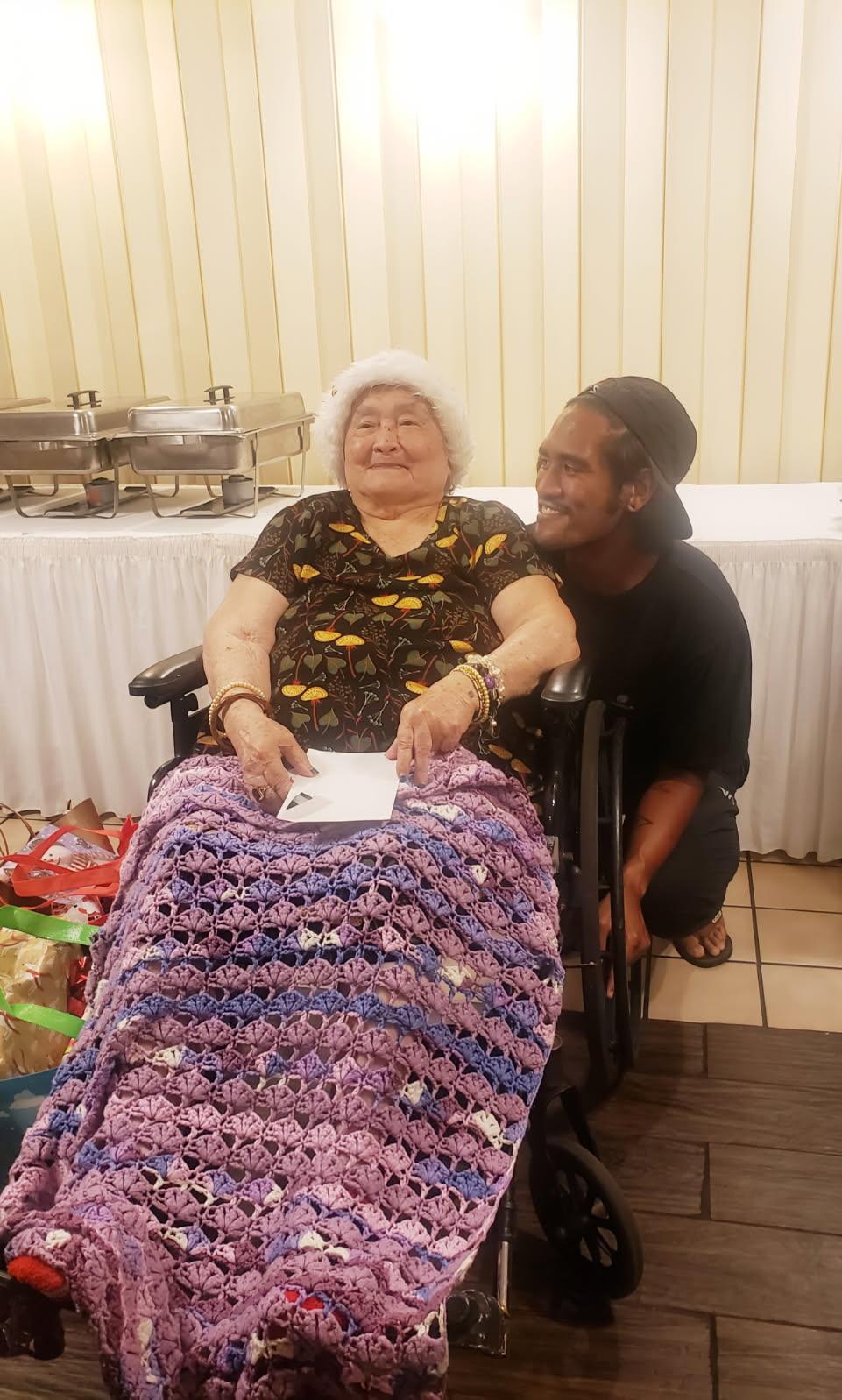 An elderly woman in a wheelchair enjoys a family gathering surrounded by gifts and loved ones.