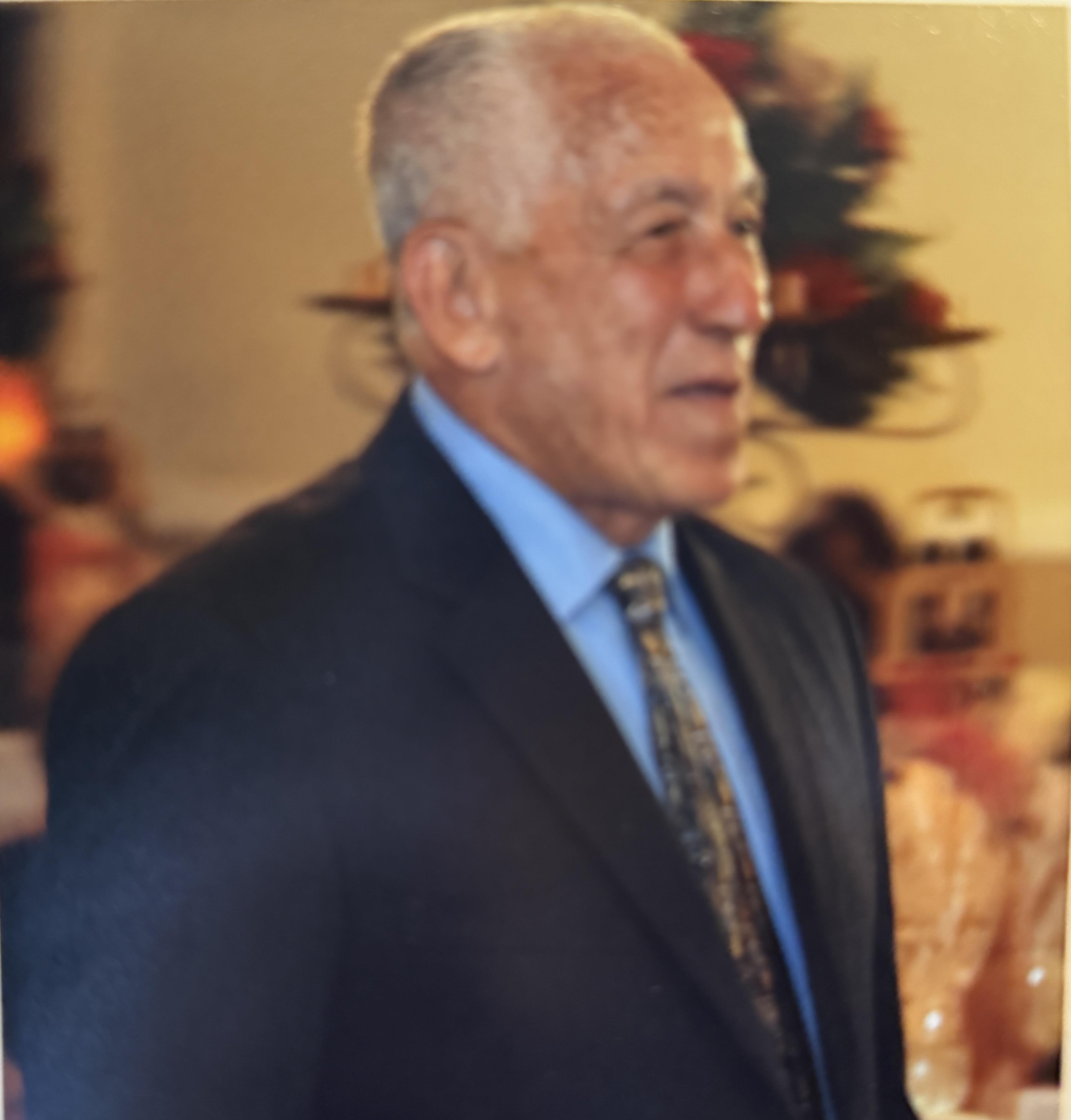 An elderly man in a suit walks through a festive venue with decorations and smiling guests.