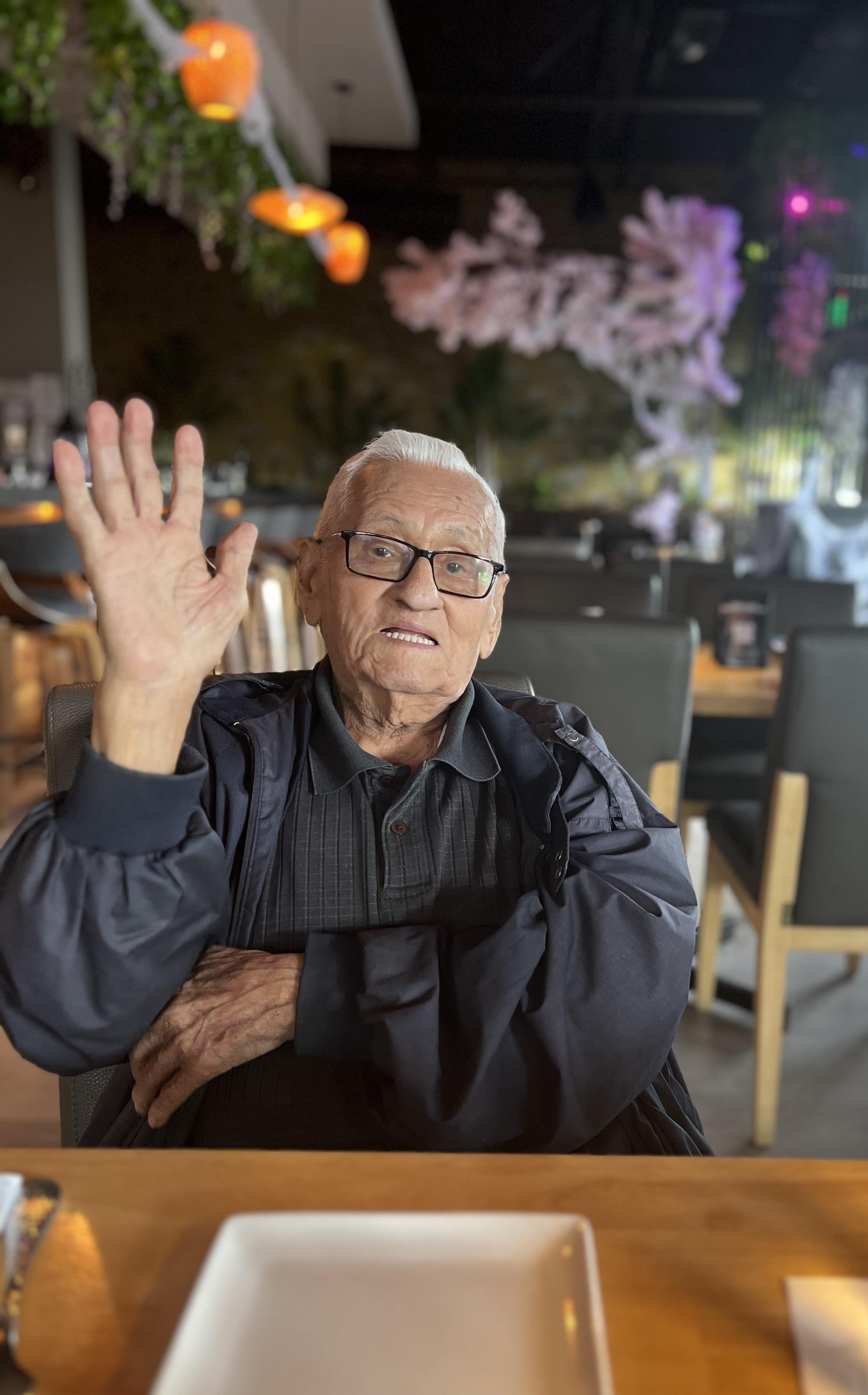 An older man with glasses raises his hand in a warm greeting while seated indoors.