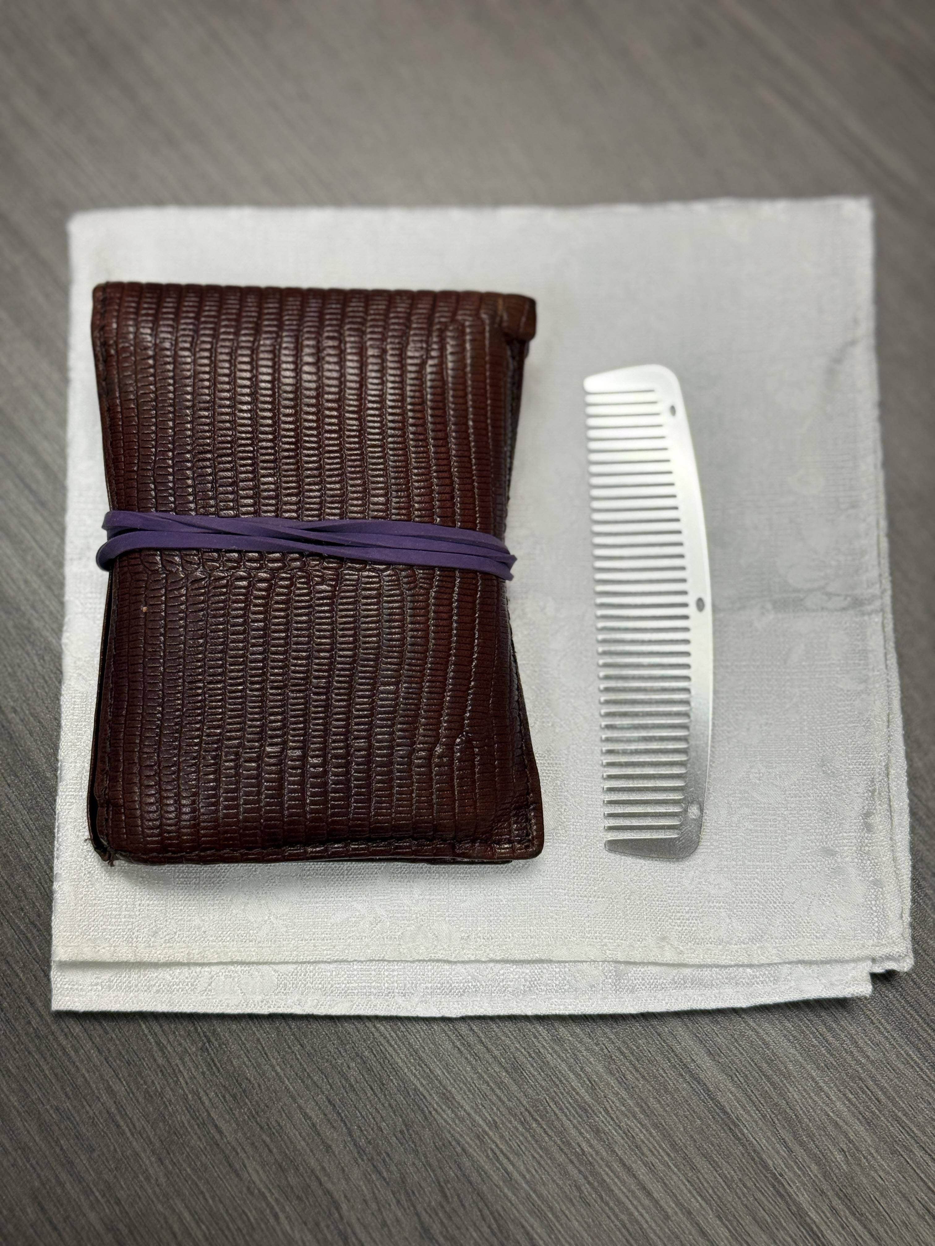 A brown wallet sits beside a silver comb on a light gray cloth.