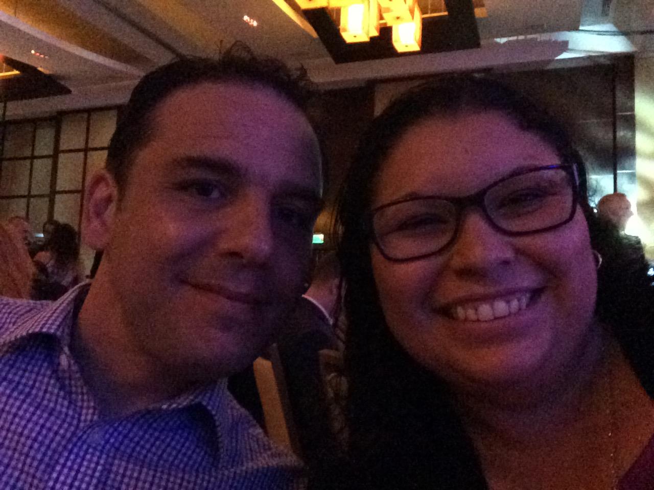 A couple smiles radiantly at the camera while attending a social gathering in the evening.