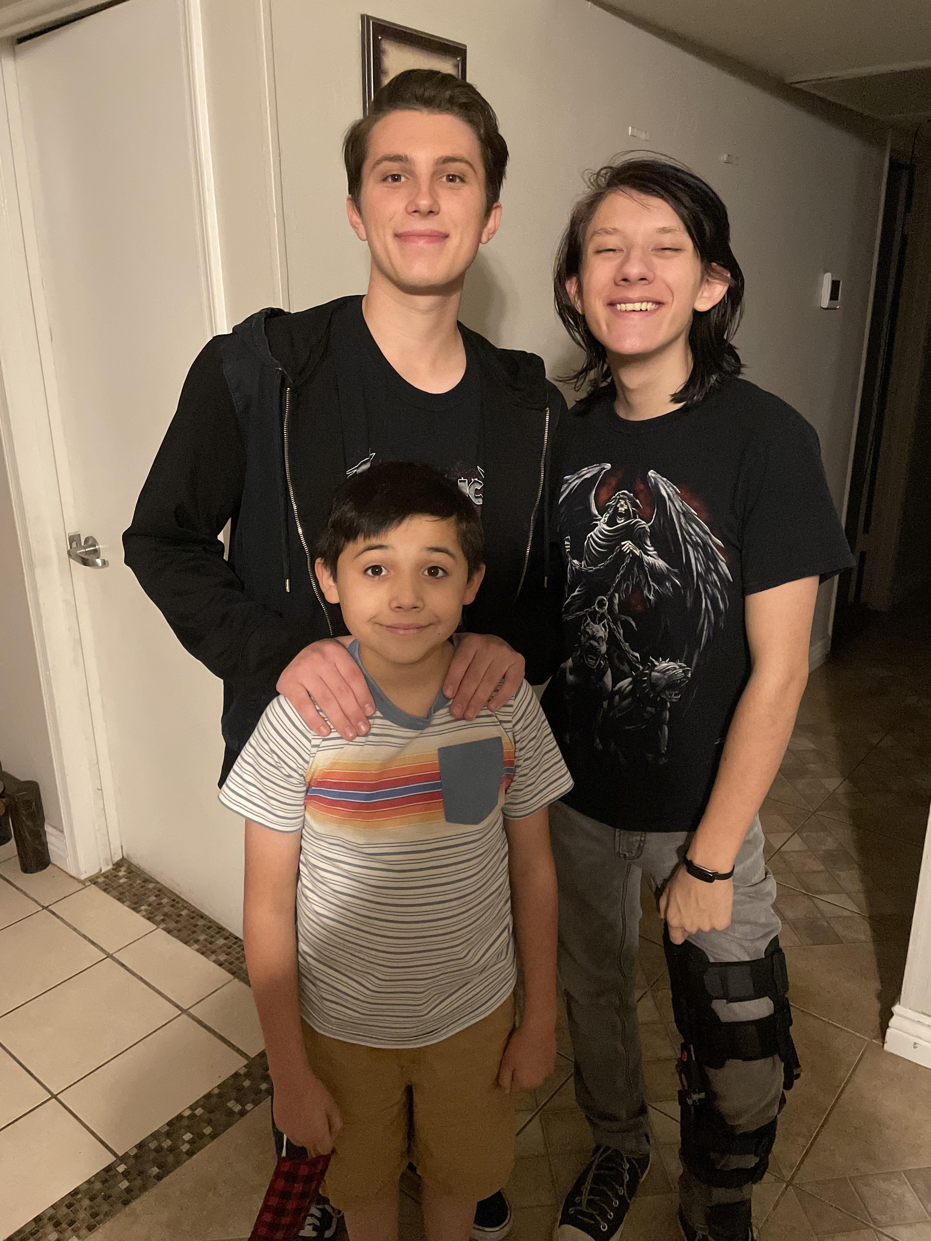 Teenagers stand and smile with a younger boy between them, enjoying a friendly moment.