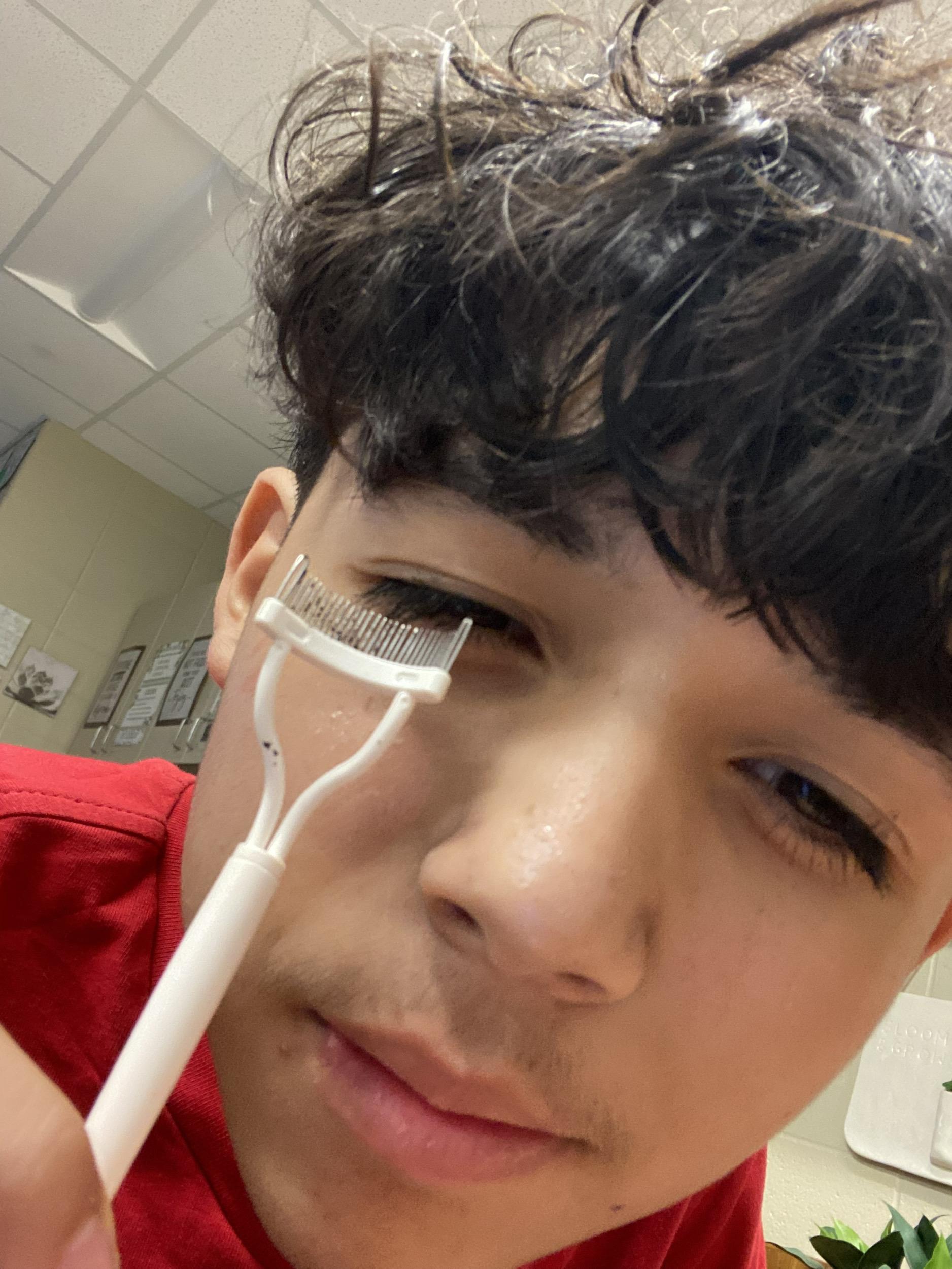 A young person holds an eyelash curler close to their eye while getting ready for the day.