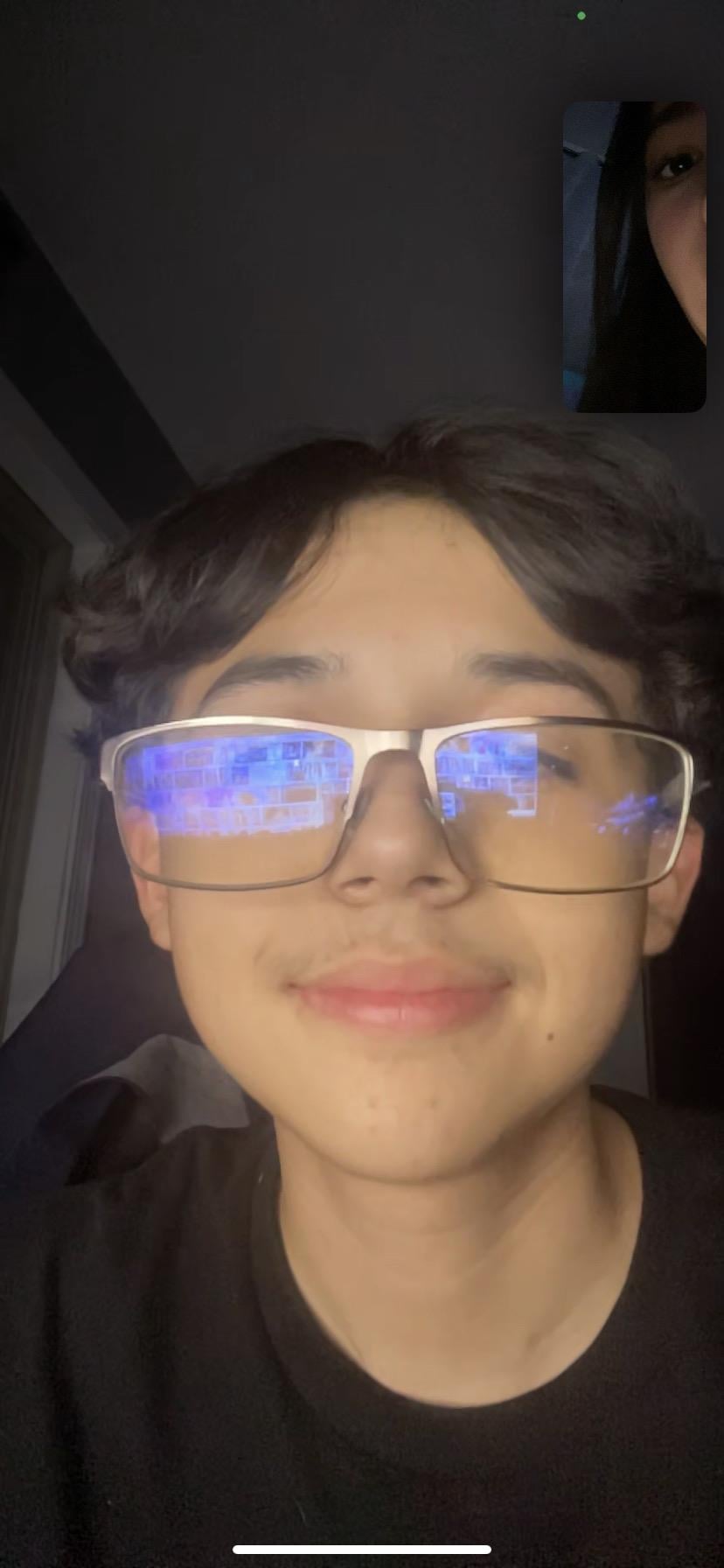 A young person looks happy with a device screen reflecting on glasses in a dark space.