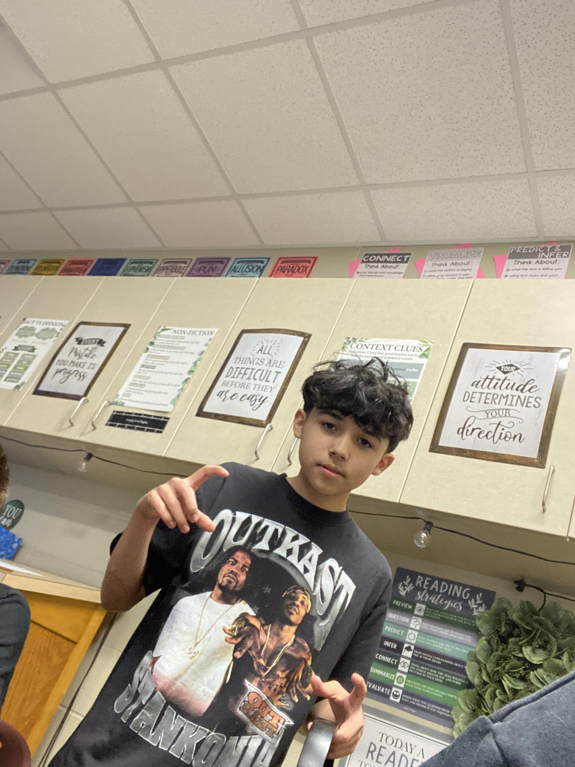 A young boy stands in front of classroom shelves while making a peace sign with one hand.