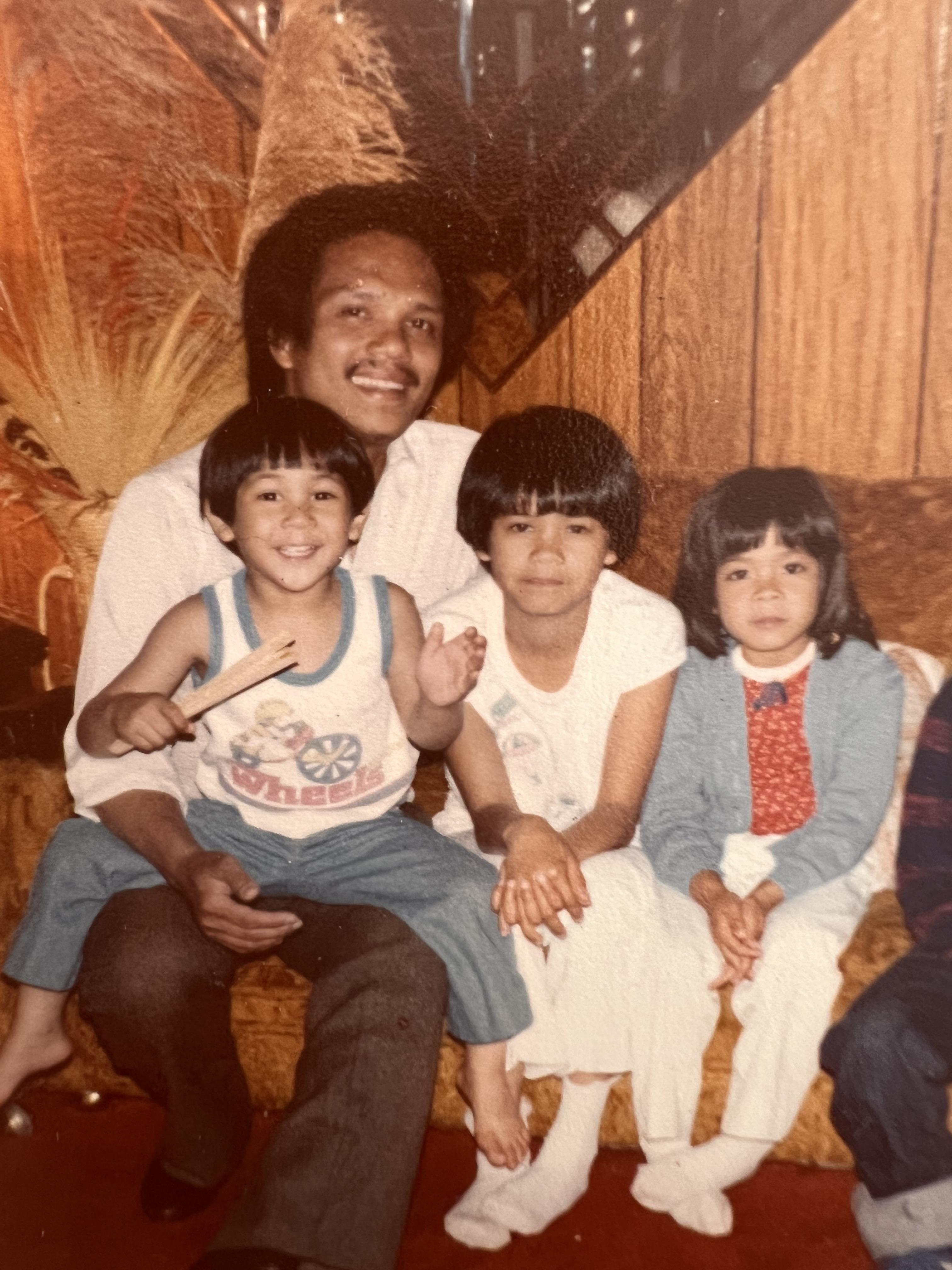 A joyful family moment shows a man with three children sitting together on a couch in the 1970s.