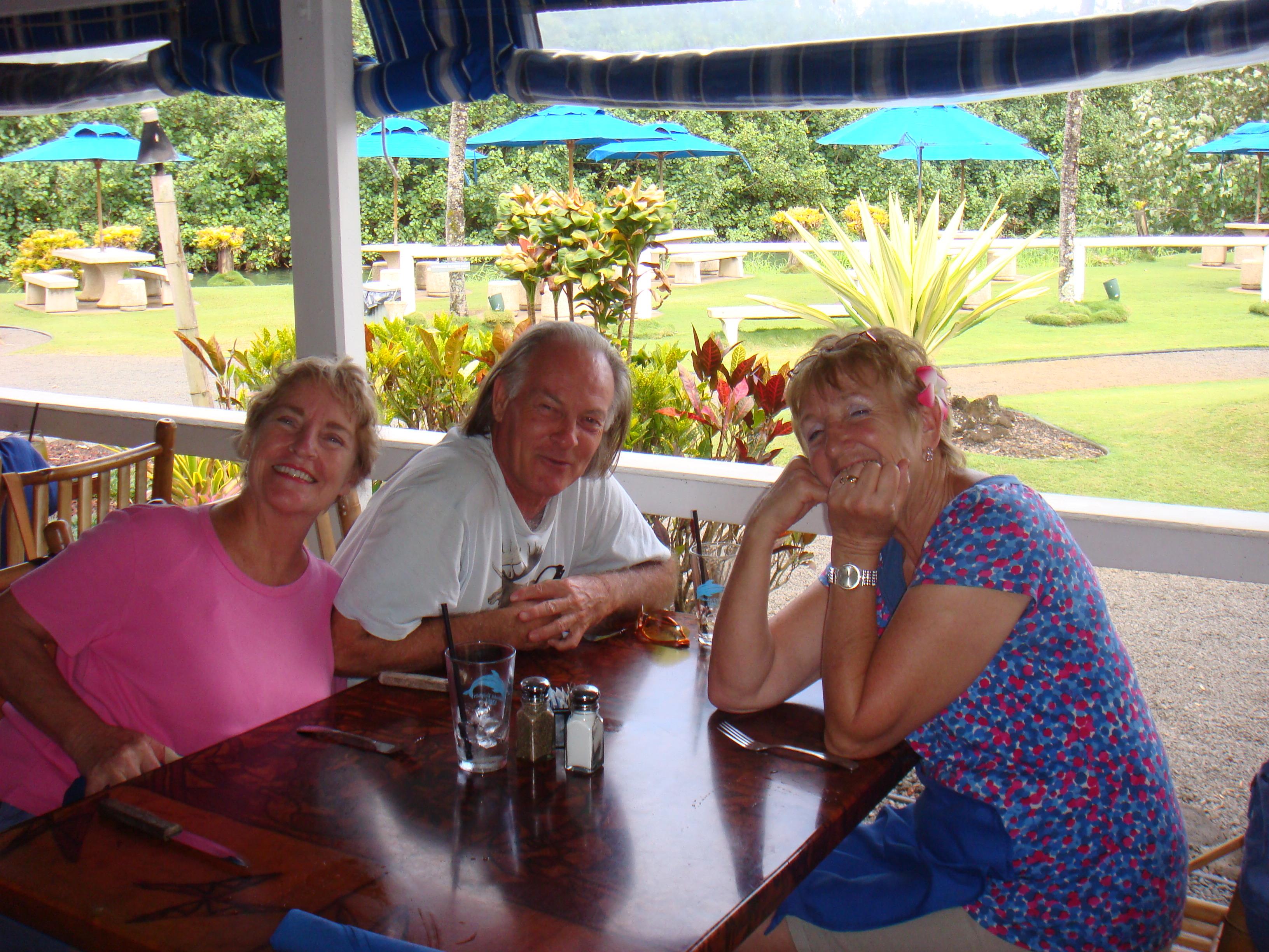 Three friends sit together at a table, sharing smiles and drinks in a scenic garden restaurant.