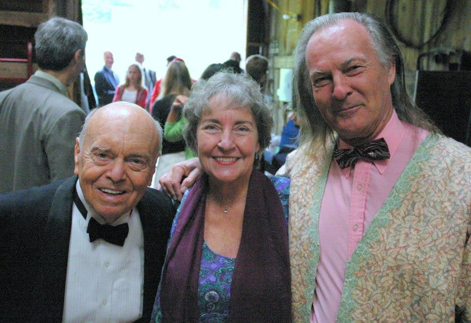 Three people in formal wear smile together at a lively event.