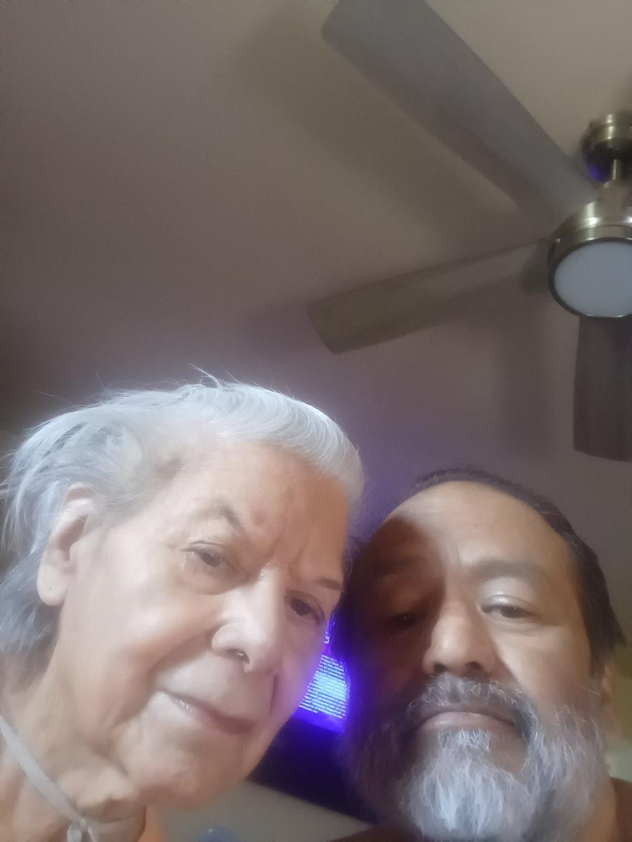 An elderly woman poses closely with a man, both smiling warmly at the camera in a cozy setting.