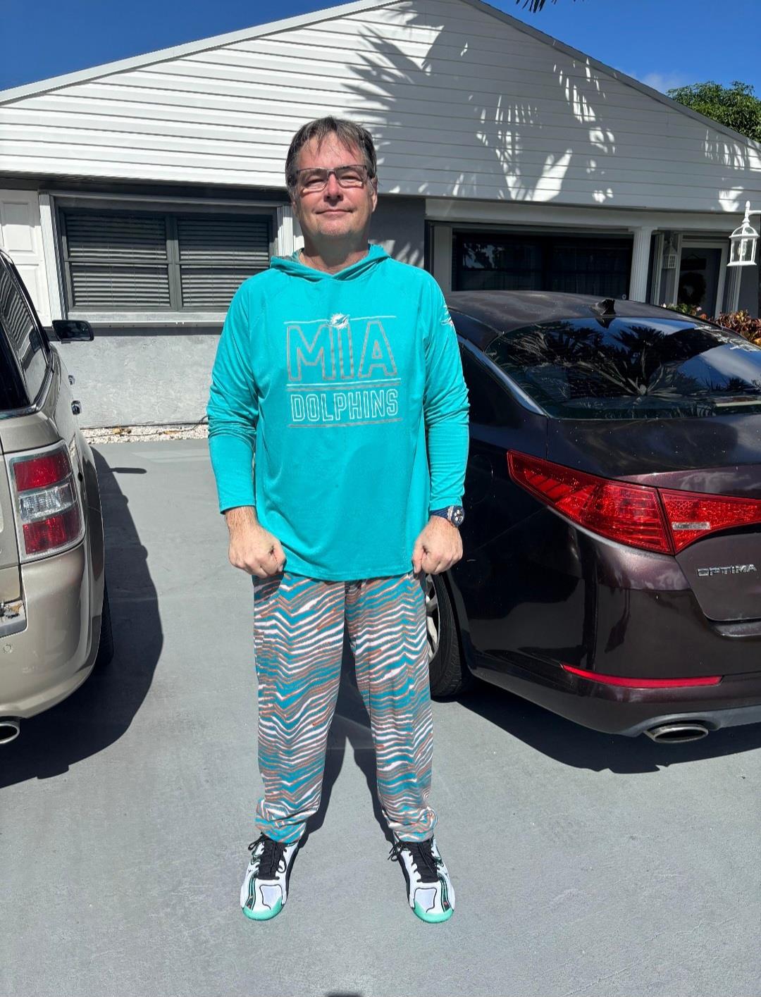 A man is smiling and posing in a vibrant outfit outside his house on a sunny day.