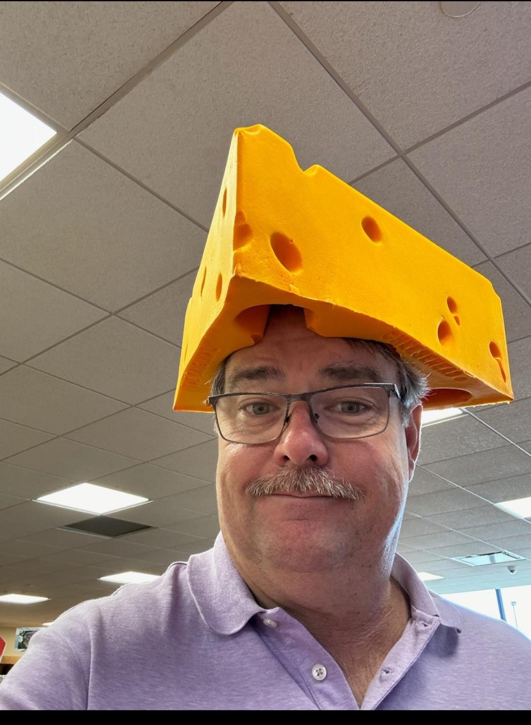 A man is smiling while wearing a bright yellow cheese hat in a lively indoor environment.