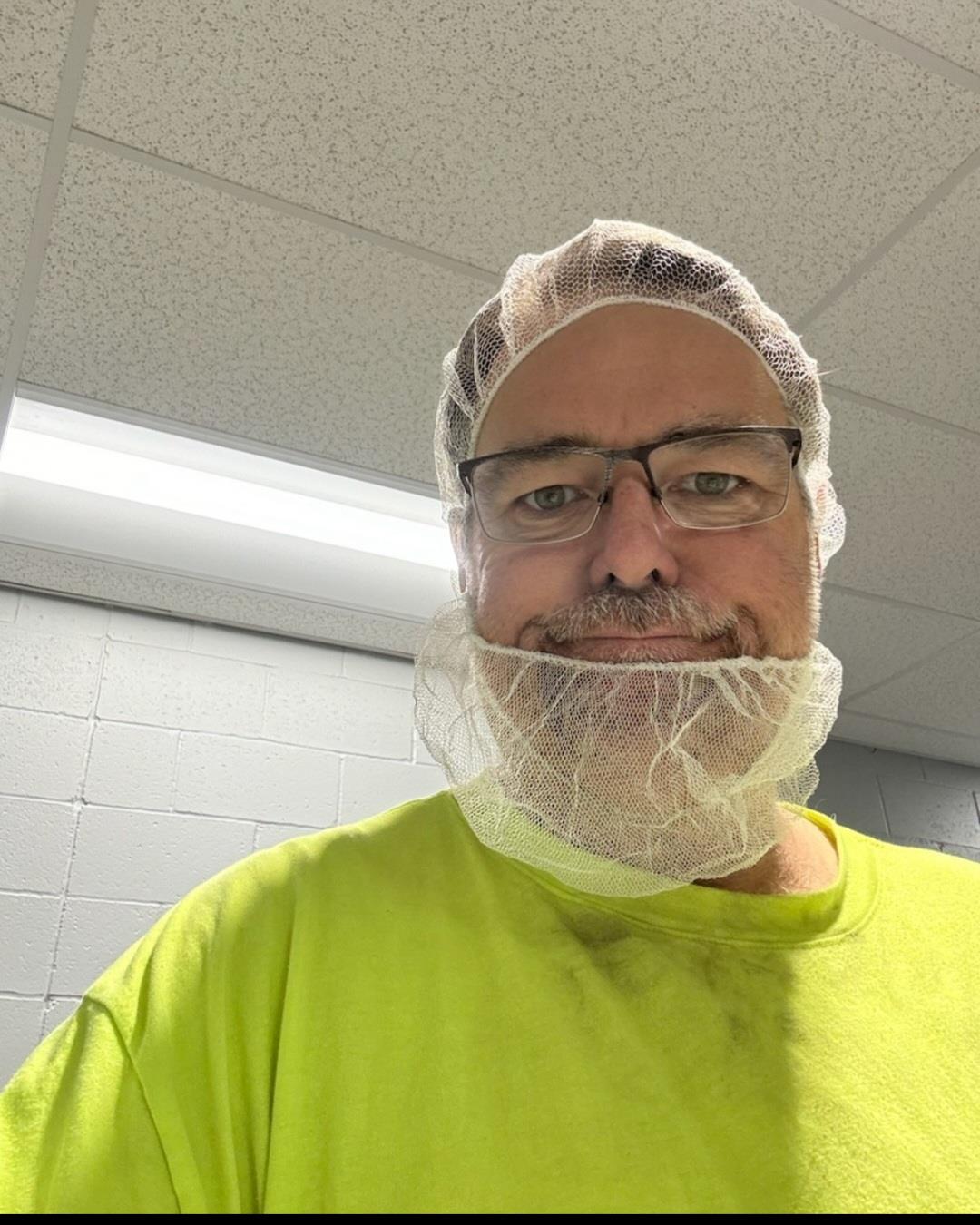 A man with glasses and a hairnet smiles while dressed in a bright shirt at a workplace.