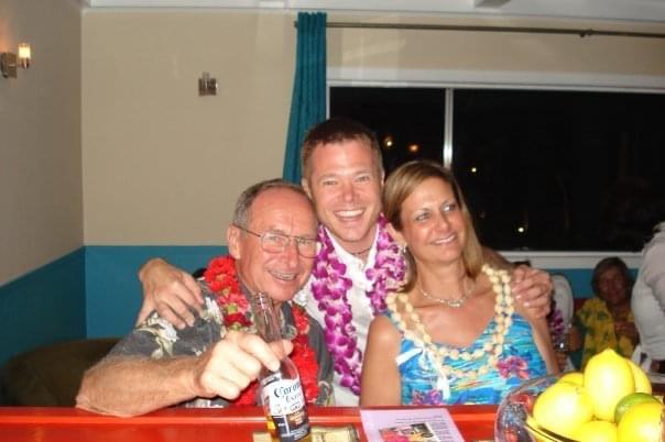 Three friends enjoy a joyful party, dressed in leis and bright attire, sharing laughter and drinks.